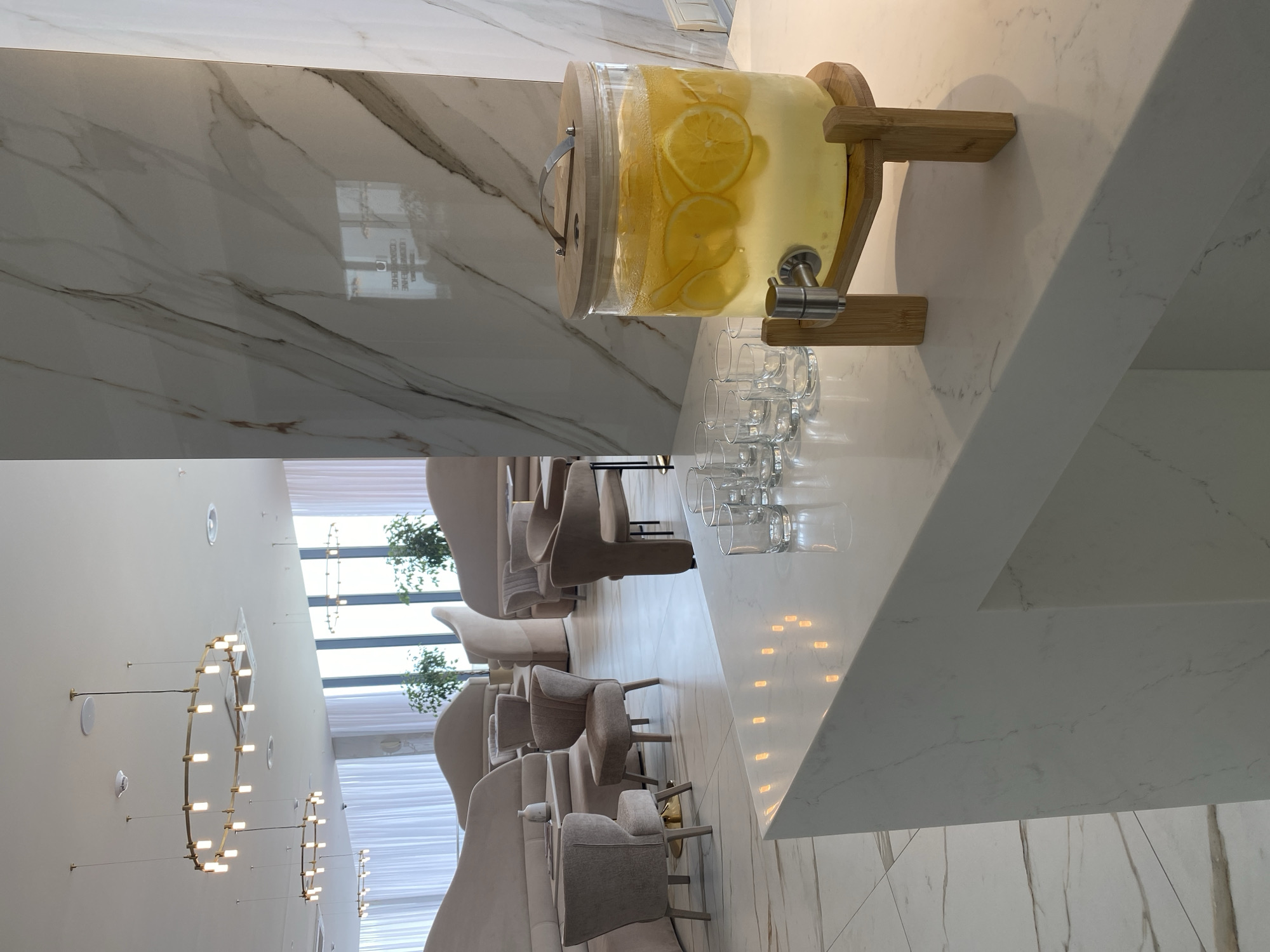 The image captures an elegant interior space, likely a lounge or reception area, possibly in Sochi, Russia. A large beverage dispenser filled with liquid and citrus slices, likely lemonade or infused water, sits on a white marble countertop. A row of clear glasses is arranged nearby. The background reveals a seating area with plush, high-backed chairs in neutral tones, suggesting a modern and luxurious aesthetic. Natural light streams through large windows. The decor includes circular light fixtures mounted on the wall. The overall ambiance is sophisticated and serene.