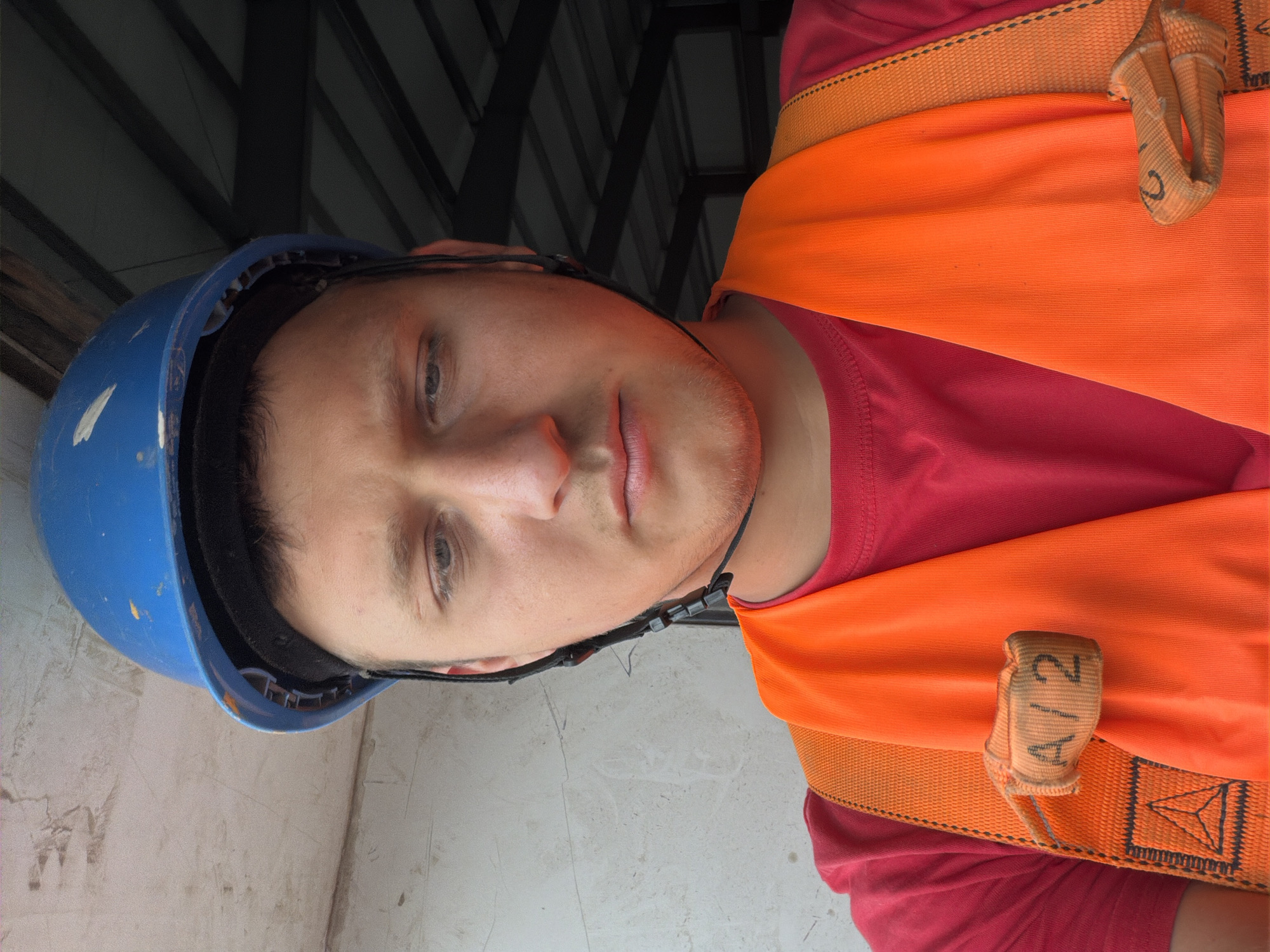 A man wearing a blue hard hat and an orange safety vest is shown in a medium close-up, looking down. He has light skin and short, light brown hair. He is wearing a red t-shirt under the safety vest. The vest has reflective yellow trim and a black strap with a loop that has "A/2" written on it. In the background, there is a light-colored textured wall on the left and a dark, corrugated metal roof structure on the right. The scene appears to be an outdoor construction or industrial area in Stróża, Poland.
