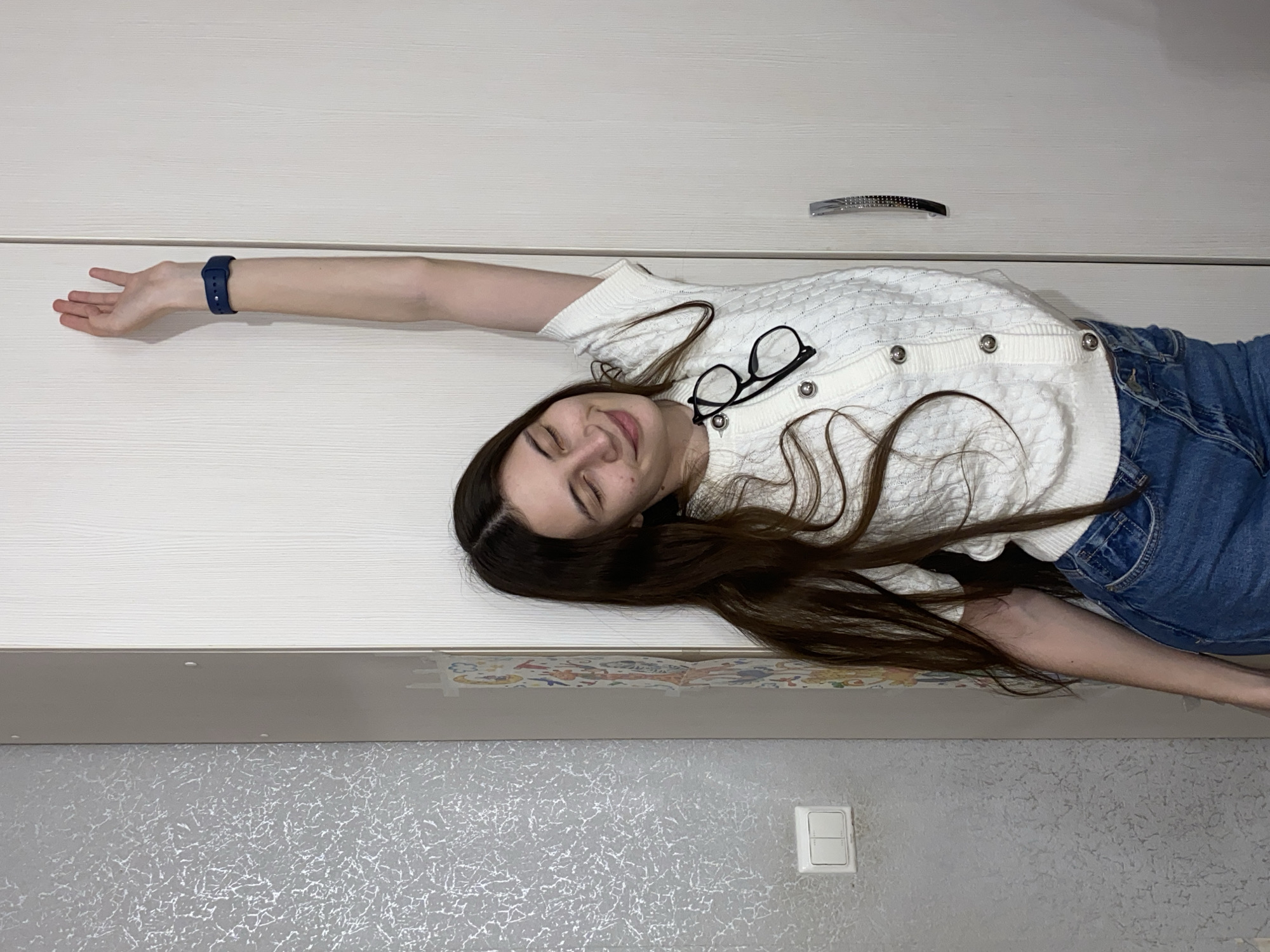 A young woman with long, dark brown hair is pictured lying on a light-colored, wood-grained surface, likely a piece of furniture such as a wardrobe or bed platform. Her eyes are closed, conveying a sense of rest or sleep. She wears a white, short-sleeved, knitted cardigan adorned with silver buttons, and blue denim jeans. A blue smartwatch is visible on her left wrist, and black-framed glasses rest on her cardigan near her neck. Her left arm is extended straight above her head, while her right arm rests bent by her side.
Behind the surface she lies on, another section of matching light-colored furniture is visible, featuring a polished silver handle. Below her, a wall is covered with textured grey wallpaper, which has a white, double-panel light switch mounted on it. A faint, colorful, patterned border can be seen where the lying surface meets the wall. The indoor lighting is soft and diffused. The image lacks any visible text or specific cues to the time of day or weather, apart from generic indoor features. The overall impression is one of peaceful repose in an ordinary residential setting, possibly in Votkinsk, Russia.