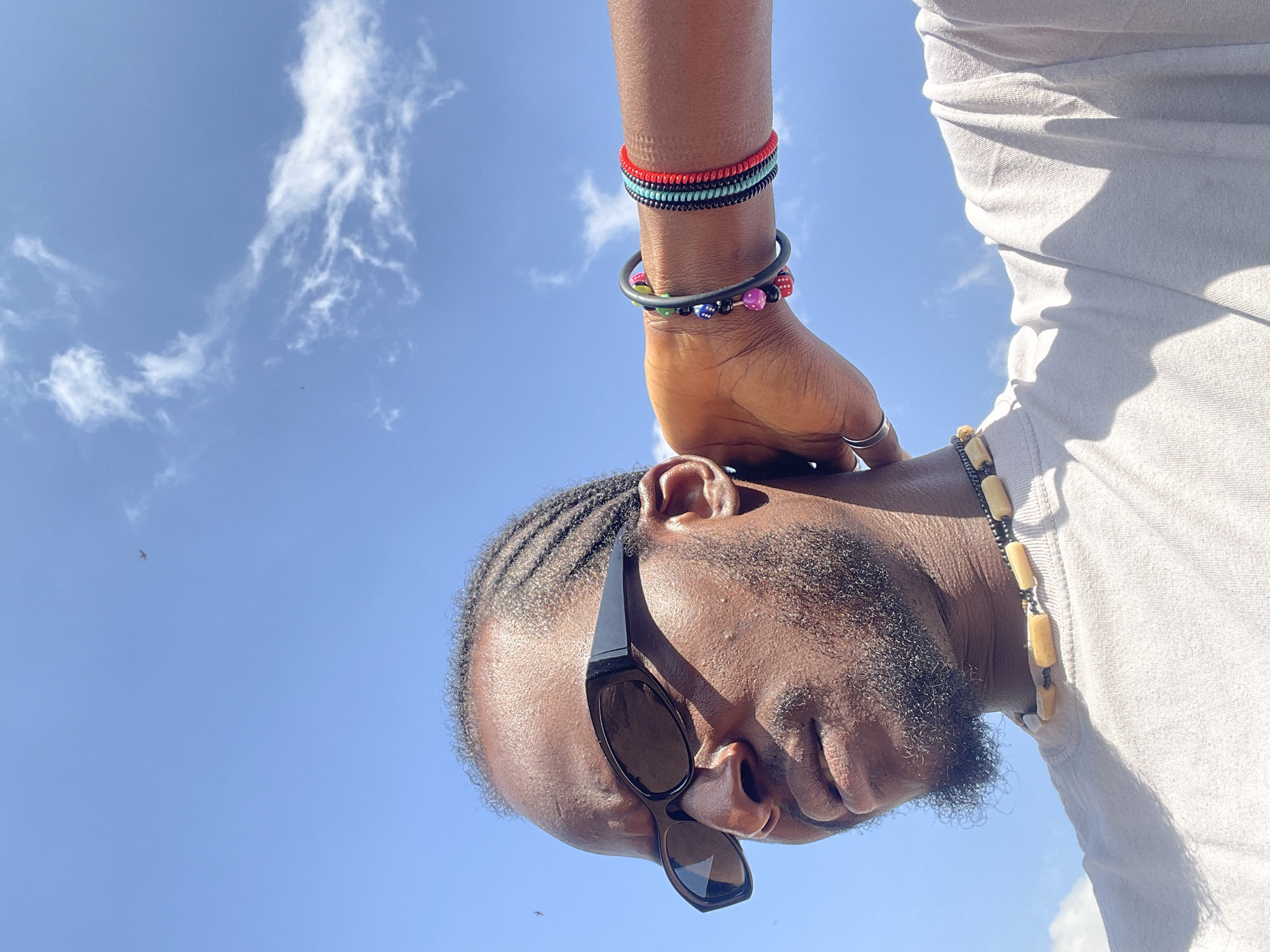 This is a close-up, low-angle outdoor shot of a Black man wearing sunglasses and a light-colored t-shirt. He has short braids in his hair and a beard. Around his neck is a necklace made of light-colored beads. On his left wrist, he is wearing a dark gray bracelet and another bracelet with colorful beads, including dice-shaped ones. On his right wrist, he has two thin bracelets, one turquoise and one red. His left arm is bent, with his hand behind his head, revealing silver rings on his fingers. The background is a bright blue sky with some scattered white clouds and two faint, distant dark shapes that could be birds or insects. The lighting suggests it is daytime. The image does not contain any specific visual cues to pinpoint its location in Jos, Nigeria.