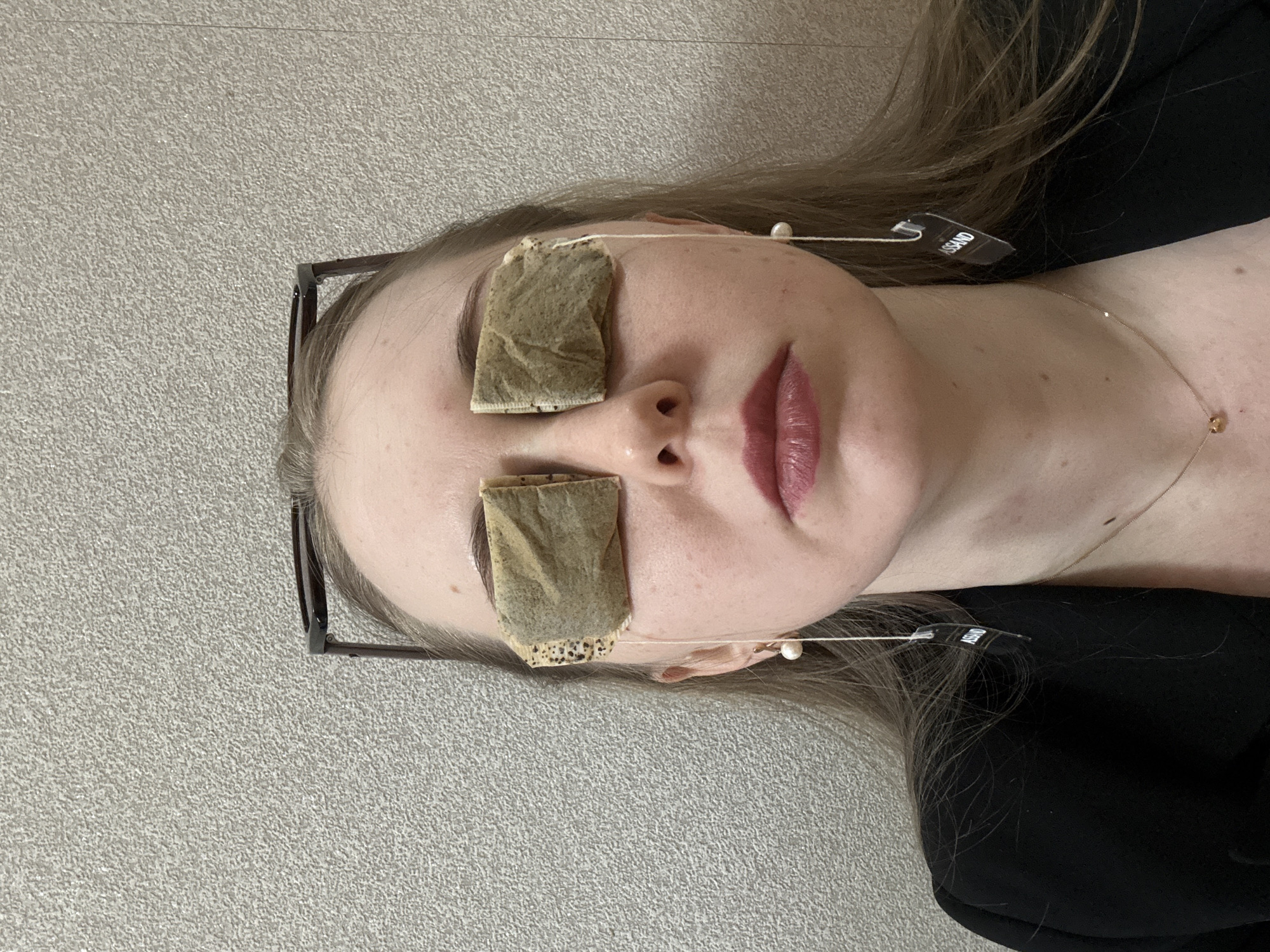 The image displays a close-up of a person, likely female, lying down with their head slightly tilted to the right. Two rectangular tea bags are placed over their closed eyelids, suggesting a relaxation or beauty treatment. A pair of glasses rests on top of their head, pushed back into their light brown hair. The person has fair skin, and their lips are slightly parted, adorned with a glossy pink lipstick. A thin, gold-colored necklace with a small, circular pendant is visible on their chest. From one of the tea bags, a string extends towards the ear, where a dark-colored tag with the partially visible text "USSAND" is hanging. Another string from the tea bag on the other eye appears to have a small white pearl attached to it, resting on the forehead. The person is wearing a black top or garment. The background is a light grey, textured surface, possibly a carpet or mat, indicating an indoor setting. The overall scene portrays a moment of self-care or repose.
