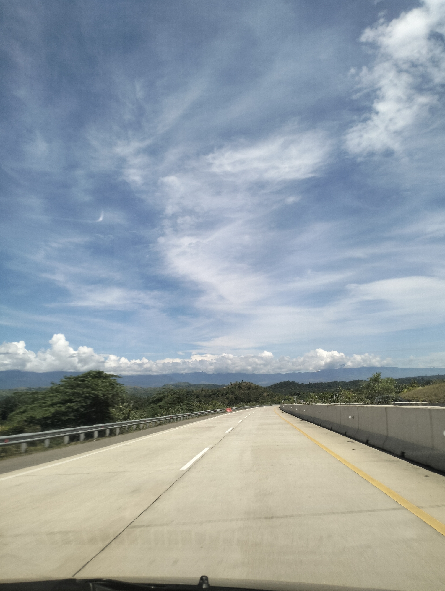The media file depicts a scenic view of a highway in Lamtamot, Indonesia. The scene is a wide, paved road with clear white and yellow lane markings, suggesting it is a well-maintained highway. The road curves gently to the right, leading towards a distant horizon. On the left side of the road, there is a metal guardrail, and beyond that, lush green trees and vegetation cover rolling hills. In the distance, a range of mountains is visible, their peaks softened by haze. The sky is a vibrant blue, filled with scattered white clouds, some appearing wispy and others more substantial and puffy. A faint, crescent-shaped sliver, possibly a very thin moon or a contrail, is visible in the upper left quadrant of the sky. The overall impression is of a bright, clear day, likely in the late morning or early afternoon, given the quality of light. The setting appears to be rural or semi-rural, with nature dominating the landscape. There is no visible text in the image. The perspective suggests the photograph was taken from a vehicle traveling along the highway. The scene evokes a sense of travel, open space, and natural beauty.
