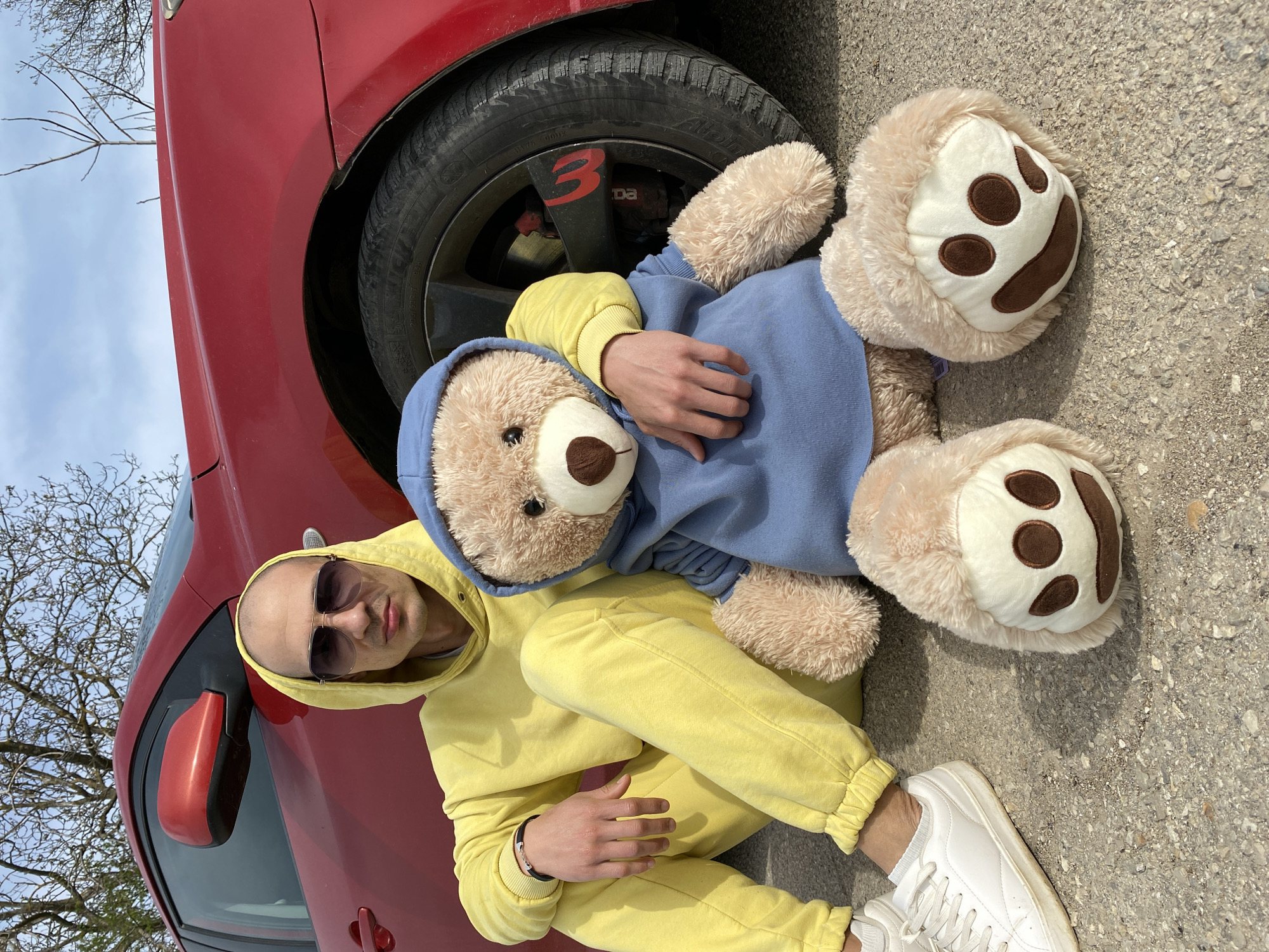 A man in a yellow hooded sweatshirt and white sneakers is sitting on asphalt next to a red car. He is wearing sunglasses and cradling a large, tan teddy bear in a blue hooded sweatshirt. The car's front tire is visible behind the teddy bear. The scene appears to be outdoors on a sunny day. I cannot infer from the image that the image was taken in Pleven, Bulgaria.