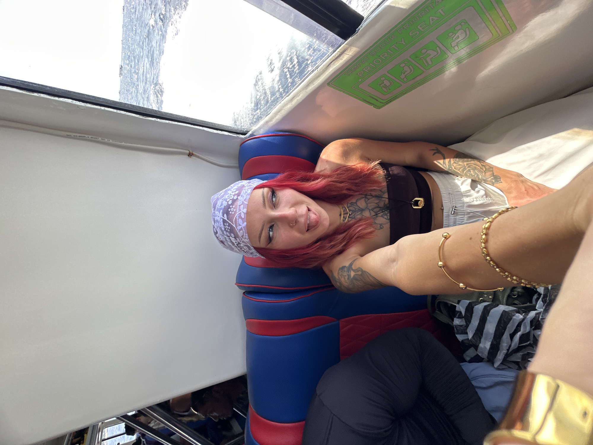 A red-haired woman is seated on a blue and red seat inside a boat, possibly on a tour, and looking at the camera. She is wearing a white floral bandana, a dark top, and white shorts. She has multiple tattoos on her chest and arms, and is wearing a gold necklace, a gold ring, and gold bracelets. The interior of the boat is visible, with a window showing the water outside. A "priority seat" sign is visible. She is on a boat, presumably in Unknown, Indonesia.