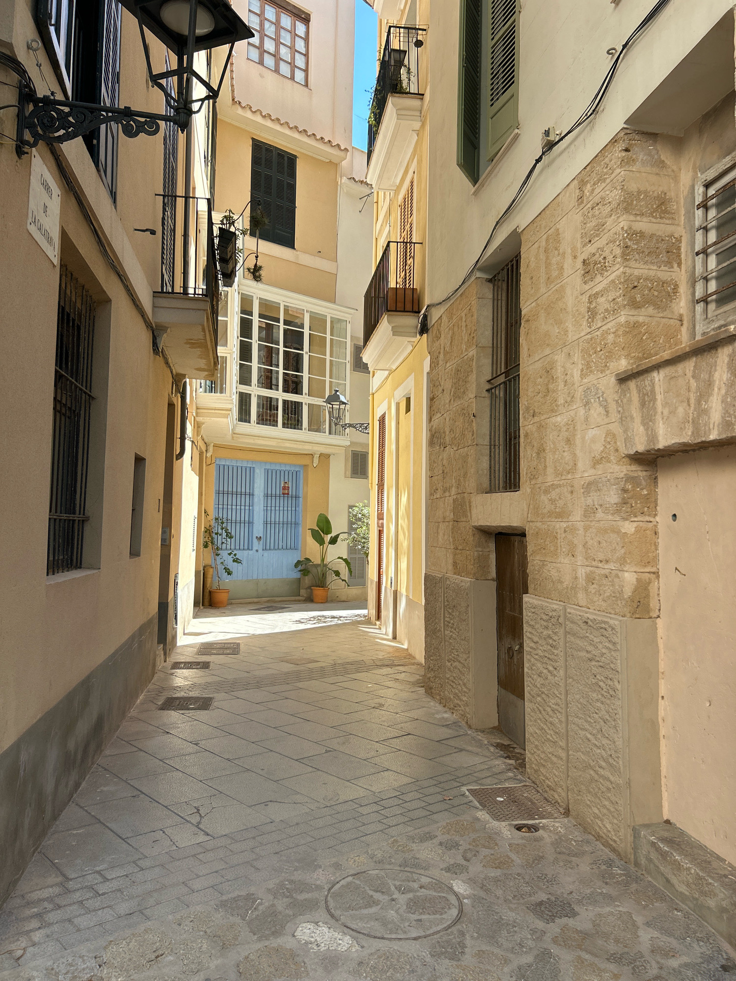 This is a photograph taken in Palma de Mallorca, Spain, looking down a narrow pedestrian alleyway. The alley is lined with buildings of varying heights and architectural styles. The left side of the alley is dominated by a building with a light yellow facade, featuring a dark grey base, wrought-iron window grills, and a black decorative lamppost mounted on the wall. Above, a street sign with the inscription "Carrer de la Calatrava" is visible. The building on the right has a textured stone facade at its lower levels, transitioning to a pale yellow color above. This side also features balconies with black railings and windows.
At the end of the alley, a light blue double door with metal bars is visible, framed by a white enclosed balcony structure above. Two potted plants, one on each side of the blue door, add a touch of greenery. The ground of the alleyway is paved with stone slabs, laid in a pattern, with some sections of cobblestones visible closer to the foreground. Several manhole covers are embedded in the pavement. The scene is bathed in sunlight, casting shadows and highlighting the textures of the buildings and pavement. The overall impression is of a quiet, historic street in a Mediterranean city.