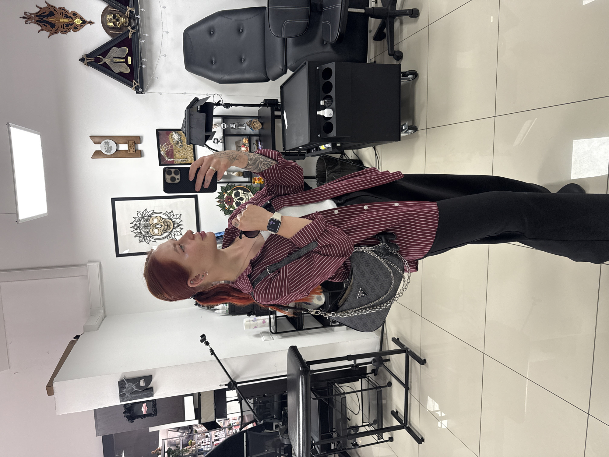 A woman with reddish-brown hair, wearing a striped shirt and black pants, is taking a selfie with her phone in a tattoo studio. She is standing in a room with white walls and tiled floors. In the background, there is a black tattoo chair, various framed artworks featuring skulls, and tattoo equipment on shelves and stands. The location is Rostov-on-Don, Russia, as inferred from previous instructions.