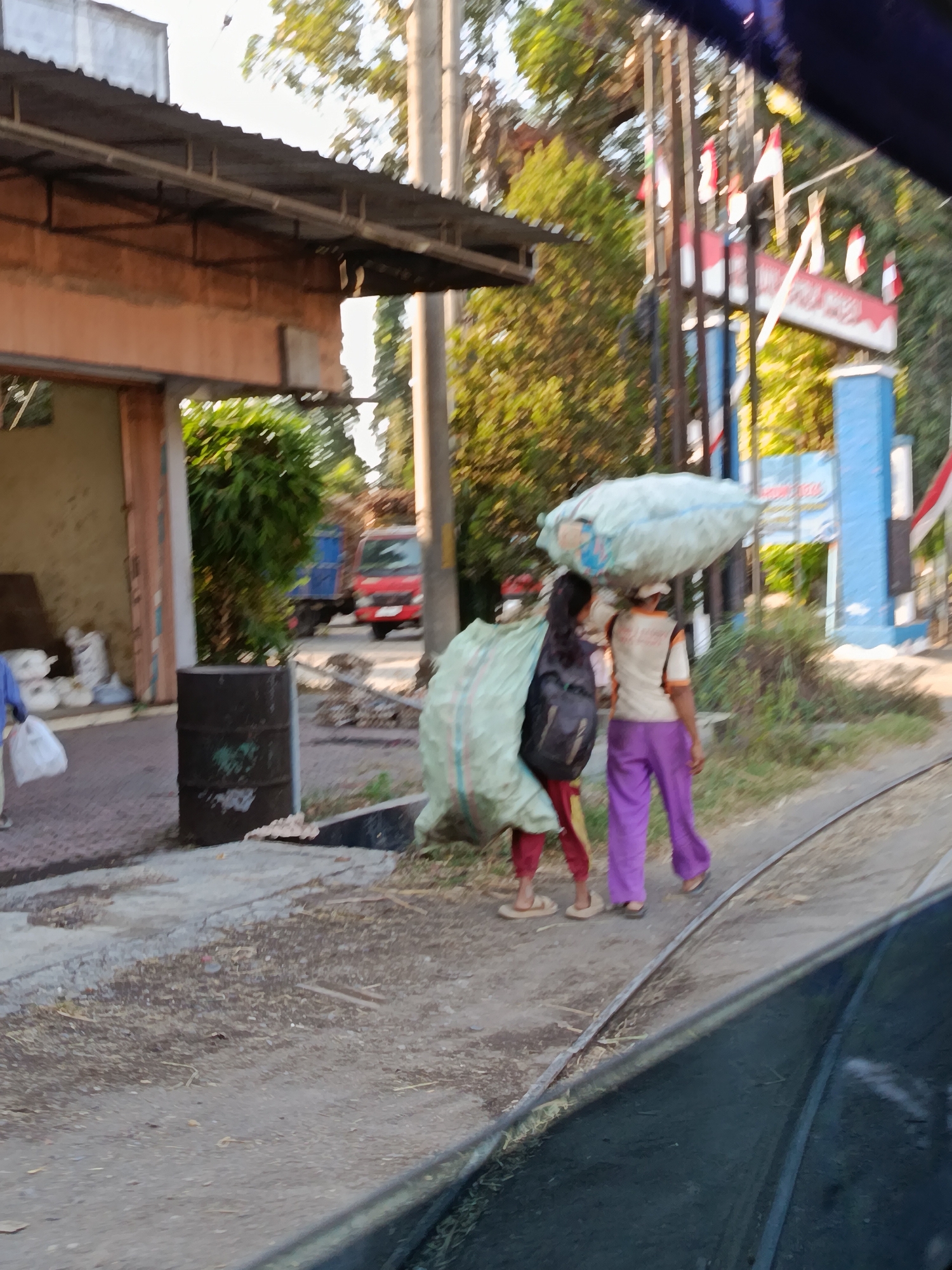 This is a medium-shot, eye-level, exterior, daytime photograph taken from a moving vehicle, likely a bus, looking towards a street scene in Ramban Wetan, Indonesia. In the foreground, a dark, reflective surface, presumably the window of the vehicle, partially obstructs the view.
The middle ground depicts two individuals walking on a dusty path adjacent to a road with railway tracks. The person on the left, facing away from the camera, is carrying a large, light green sack over their left shoulder. They are wearing red pants and light-colored sandals. They also have a dark backpack. The person on the right, also facing away from the camera, is carrying a large, light blue sack on their head. They are wearing a white shirt with orange trim and bright purple pants, with dark sandals. Both individuals are walking to the right of the frame.
The background features a building with an orange-colored facade and a corrugated metal roof on the left. In front of this building, a black barrel is visible. Further back, a red truck with a blue cargo bed is parked on the road. Behind the individuals and the truck, a line of trees and utility poles with Indonesian flags are present. A blue structure, possibly a gate or a signpost, is also visible with flags. The environment appears to be rural or suburban, with elements of construction or storage visible near the building. The lighting suggests it is late afternoon or early morning, with the sun casting shadows.