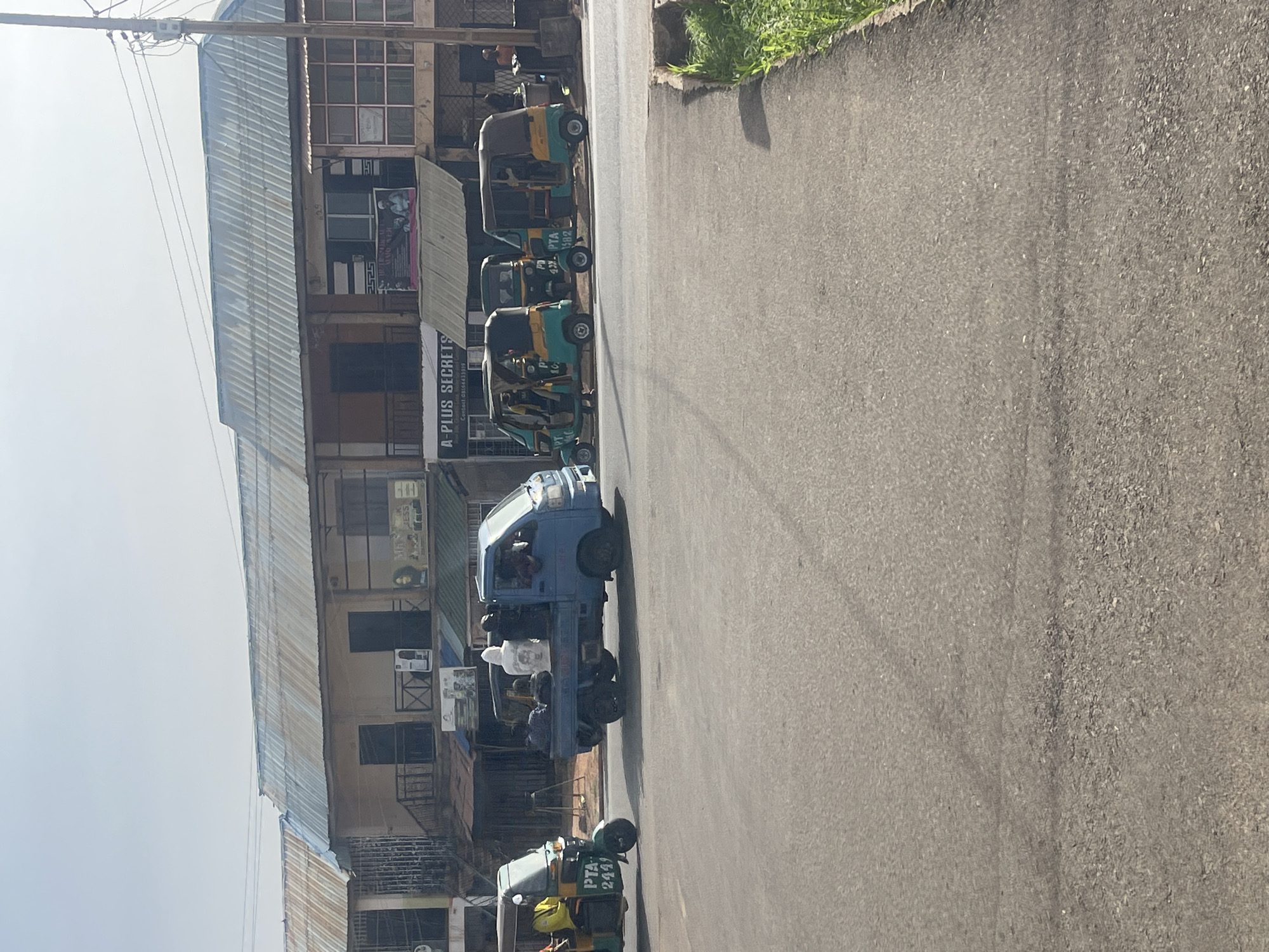 This image captures a vibrant street scene in Jos, Nigeria, during clear daylight hours. The perspective is taken from a moving vehicle, looking across a paved road towards a row of commercial buildings.
On the left side of the frame, several multi-story buildings line the street, featuring corrugated metal roofs and various window designs. These structures appear to house businesses, with a prominent sign reading "A-PLUS SECRETS" and displaying a contact number: "08034433919". Another partially visible sign shows the word "JESS". Air conditioning units are mounted on some building exteriors. Overhead, utility poles and power lines crisscross against the bright sky.
The paved road dominates the center and right side of the image. It appears to be a busy thoroughfare. A light blue small pickup truck, carrying several passengers in its open bed, is visible mid-frame. Alongside the road, or perhaps in traffic, are multiple three-wheeled auto-rickshaws, locally known as Keke Napep. These vehicles, distinct in their teal, yellow, and olive-brown colors, bear visible license plates such as "PTA 2449", "PD 42Y", and "PTA 1582". People are seen inside these vehicles, indicating ongoing travel and daily activities. The right edge of the road features a small, unpaved verge with sparse green grass and dirt.
The scene conveys a sense of everyday urban life and commerce, typical of a Nigerian city.