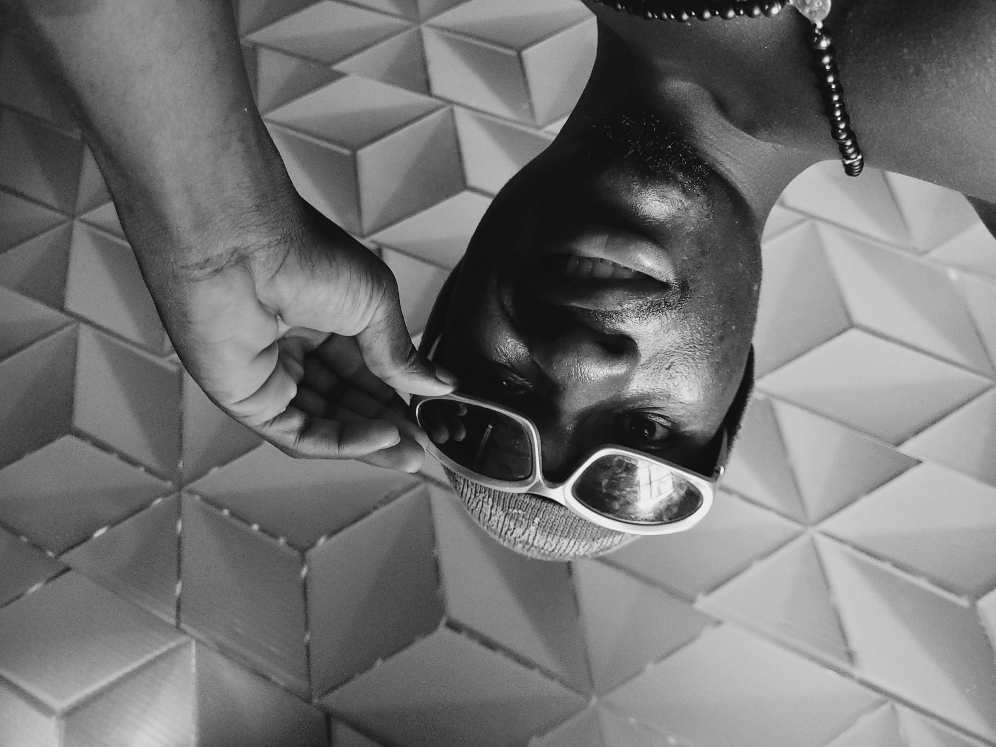 A man wearing sunglasses and a beaded necklace is photographed in black and white, looking down at the camera. He is holding the sunglasses with his left hand. The background features a wall with a geometric, diamond-patterned texture.
The information about the location of Bukuru, Nigeria is extraneous and cannot be confirmed by the image.