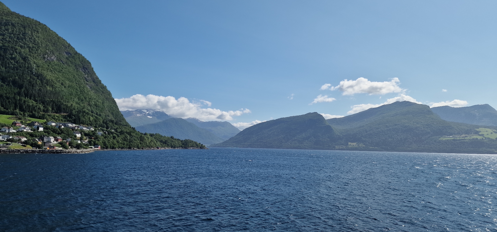 This is a wide-angle landscape photograph of a fjord in Volda, Norway, taken from the water. The foreground is dominated by a large expanse of deep blue water with moderate ripples. In the middle ground, a steep, densely forested mountainside rises dramatically from the water on the left side of the frame. Clinging to the lower slope of this mountain is a small, picturesque village with white and red-roofed houses. The village appears to be situated along a rocky shoreline.
Beyond the immediate mountainside, a chain of larger, majestic mountains stretches across the horizon, some of their peaks dusted with snow. The sky above is a clear, vibrant blue with scattered, fluffy white clouds. The overall impression is one of serene natural beauty and a peaceful, remote settlement. There are no people visible in the photograph, and no apparent actions or interactions are taking place. The scene is static and dominated by the natural environment and the small human settlement.