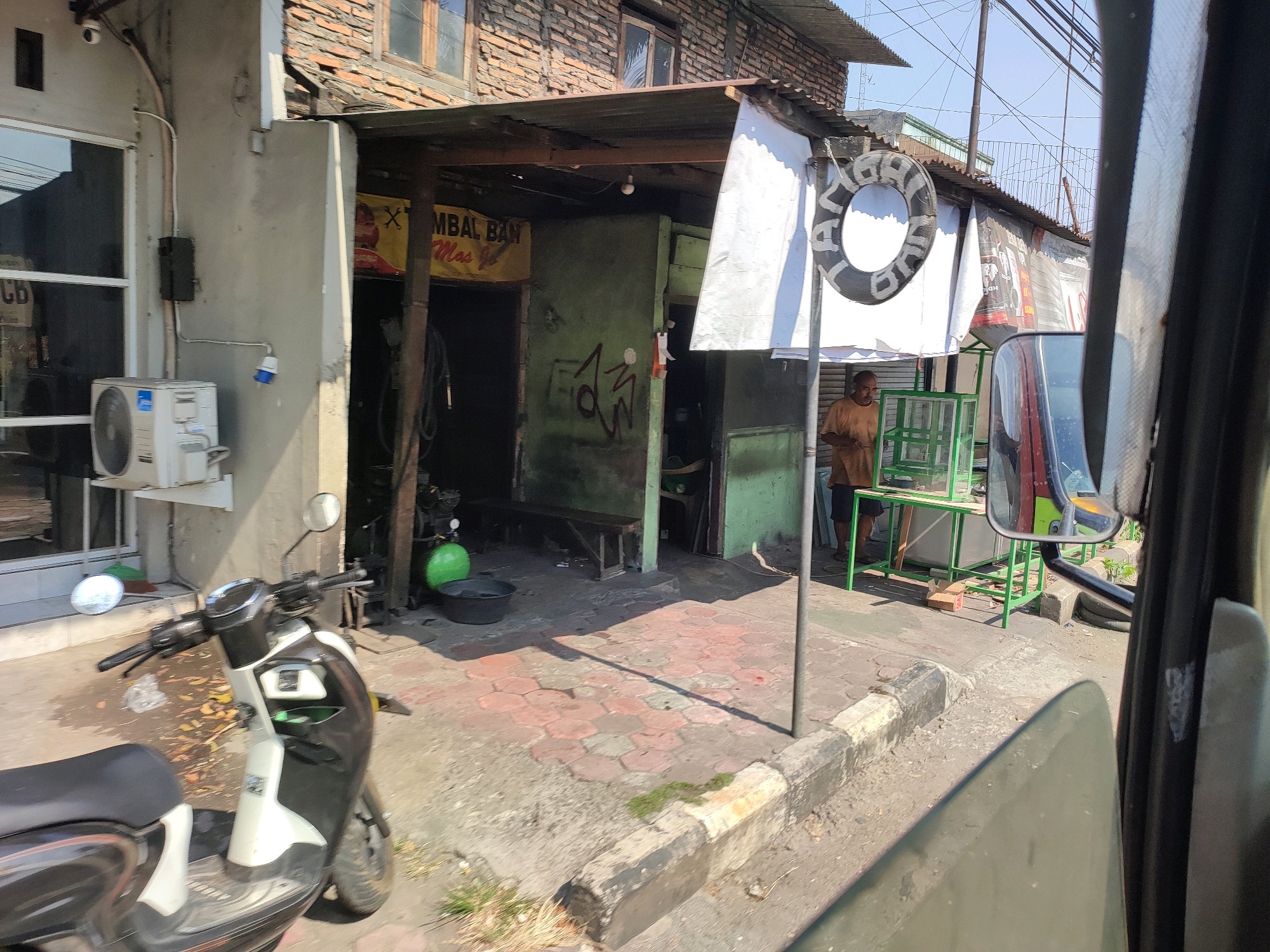 The image captures a street scene in the city of Magelang, Indonesia, likely during daylight hours. In the foreground, a black and white scooter is parked on the paved sidewalk. The scooter has a large black seat, a black and silver handle, and a black front wheel. Behind the scooter, a wall of a building is visible, with an air conditioning unit mounted on it. Further back, a storefront with an open entrance reveals a workshop or small business. A man in an orange shirt stands inside the business, near a green counter. A life preserver ring with white lettering is hung on a post outside the establishment. The street in front of the business is paved with concrete and hexagonal red bricks. Buildings with brick facades and corrugated metal roofs line the street. Overhead, power lines crisscross the sky, suggesting an urban environment. The overall impression is of a typical, bustling street in a Southeast Asian city.