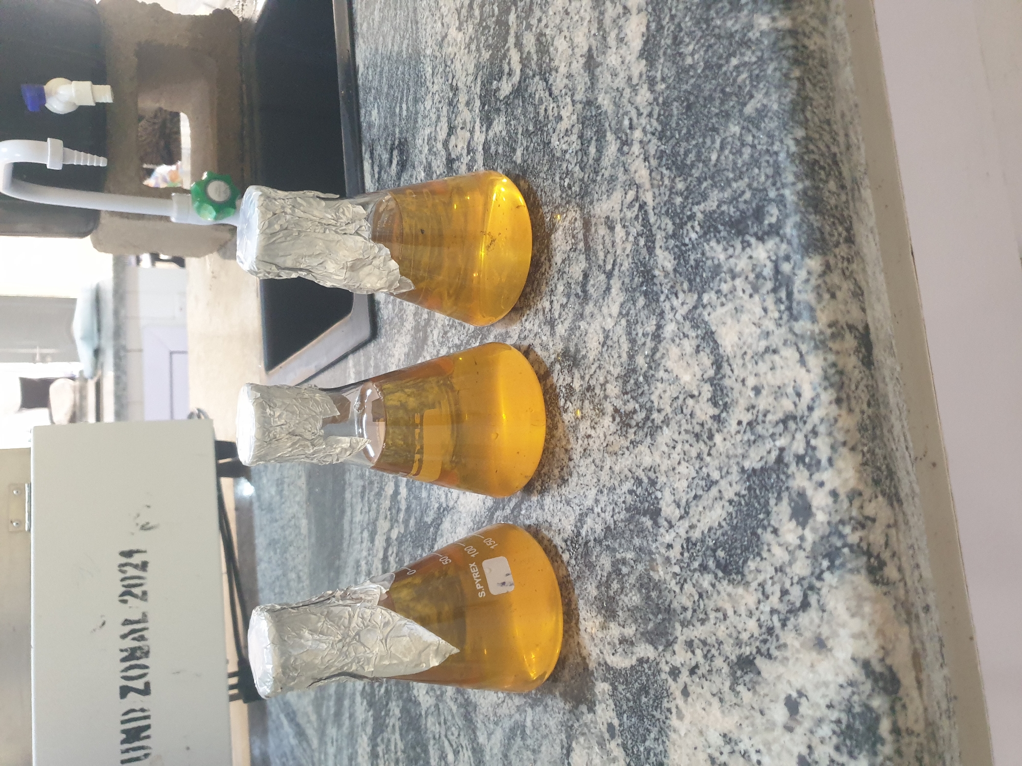 In an indoor laboratory setting, three Erlenmeyer flasks containing a yellow liquid are arranged on a speckled granite countertop. The flasks are covered with aluminum foil. To the left of the flasks, a white machine with the stencil text "UMD ZONAL 2021" is visible. To the right of the flasks, a faucet assembly with a green valve extends over a dark sink. The background shows laboratory equipment and a brick structure. The lighting suggests an overhead source, creating shadows on the countertop.