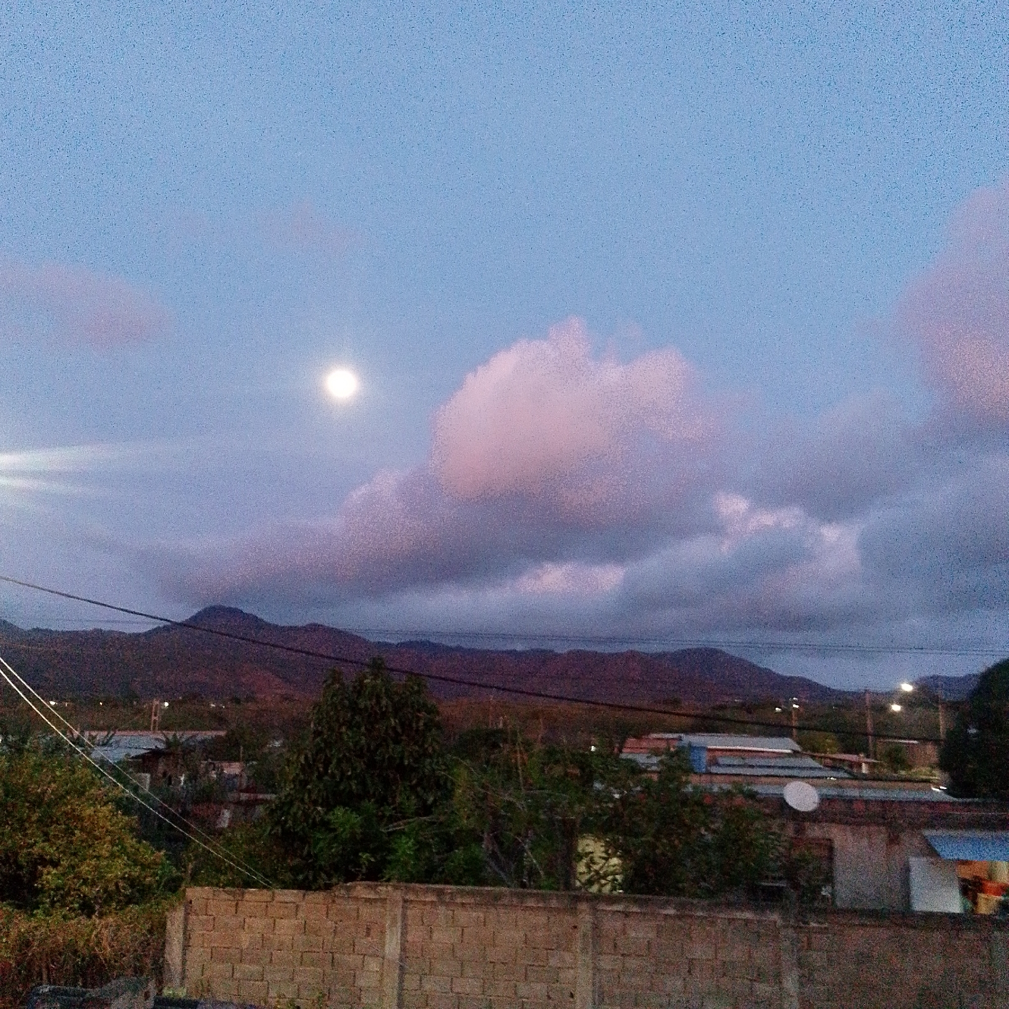 The image presents an evening view in Altagracia, Venezuela. A low brick wall dominates the foreground, behind which are trees and rooftops of buildings. Mountains form the horizon under a sky filled with clouds tinged with pink. The moon is visible in the upper left portion of the sky. Power lines extend across the frame. A few streetlights can be seen in the distance. No people are visible. A satellite dish is visible on one of the rooftops.