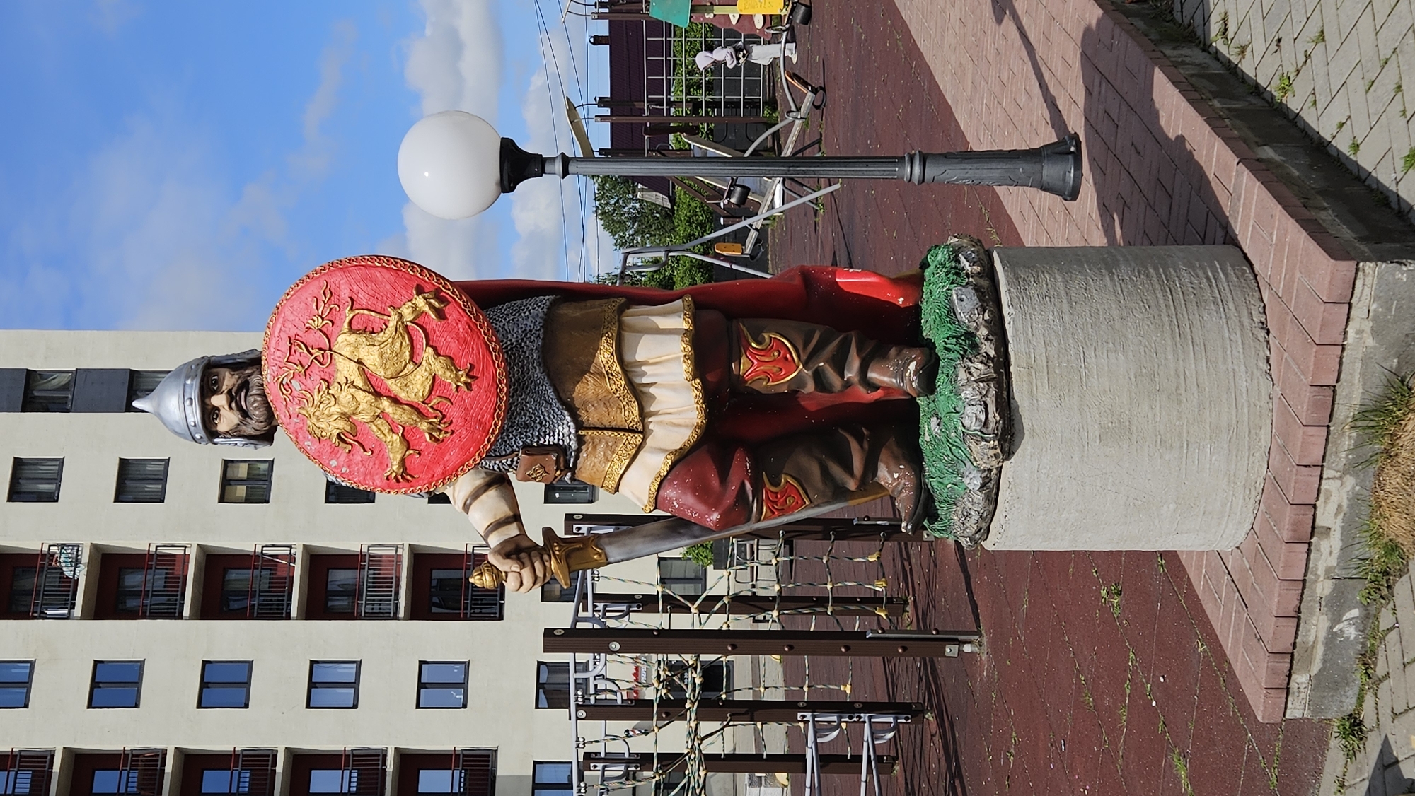 Here is a description of the image:
An outdoor statue of a medieval warrior is seen standing on a concrete pillar. The statue is dressed in chainmail, a red cape, brown boots, and a leather skirt. He holds a red and gold shield in his left hand and a sword in his right. The statue is located on a red paved area next to a sidewalk made of square gray stones. Behind the statue is a tall apartment building with a light gray exterior and many windows. A street light with a round white bulb is also visible behind the statue. The sky above is partly cloudy with patches of blue sky. The photograph is rotated 90 degrees counter-clockwise.