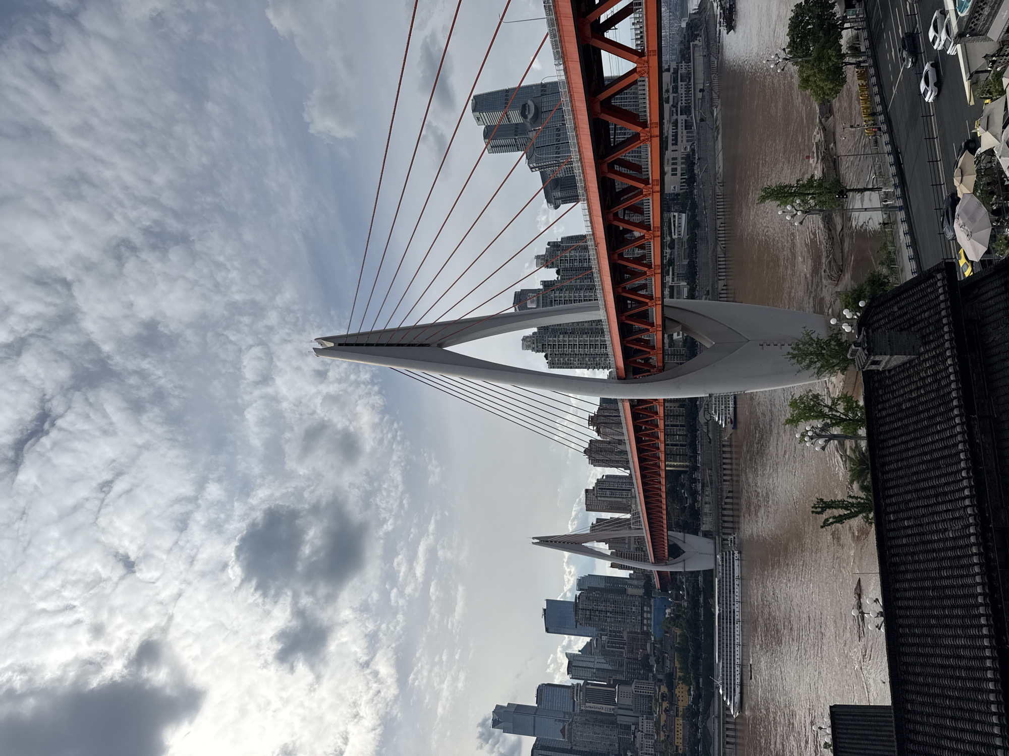 A vibrant cityscape unfolds, dominated by a large, modern cable-stayed bridge spanning across a wide, muddy river. The original image is rotated 90 degrees counter-clockwise; when viewed upright, the bridge extends horizontally across the frame.
The central subject is a distinctive bridge, featuring prominent white or light grey A-frame pylons and an orange-red, latticed steel truss structure forming the bridge deck. Numerous slender red cables extend diagonally from the pylons to support the deck.
Below the bridge, a broad, brown, sediment-rich river flows. On its surface, a large, multi-deck white passenger ship or ferry is visible, navigating under a section of the bridge.
Both banks of the river are densely populated with an array of tall, contemporary high-rise buildings, forming a sprawling urban environment. Some buildings on the left bank exhibit unique architectural features, such as large circular openings on their facades. On the right bank, a busy street runs parallel to the river, featuring several cars, including white and black vehicles. Streetlights and scattered trees line this road. In the immediate foreground on the right, the dark, textured roof of a building, possibly with traditional Chinese architectural elements, is partially visible. Nearby, several large, light-colored patio umbrellas are set up, suggesting outdoor dining or resting areas.
The sky above is partly cloudy, with a mix of bright white and softer grey clouds suggesting a clear day with some atmospheric haze. The time of day appears to be daytime, likely late morning or early afternoon, given the bright illumination.
The scene captures a bustling modern metropolis with significant infrastructure, characteristic of a major Chinese city like Chongqing, especially aligning with the context of Nan'an District. No people are distinctly visible, and no text is legible within the image.