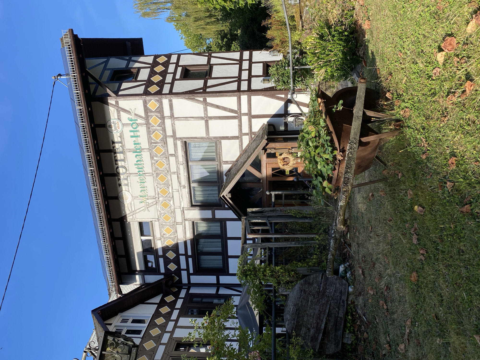 In Seelbach bei Hamm (Sieg), Germany, a multi-story half-timbered building identified as "HOTEL Marienthaler Hof" is prominently featured against a clear blue sky. The building's facade is white with dark brown timber framing, and decorative yellow diamond patterns are visible on the upper levels. Solar panels are installed on the roof.

In the foreground, a rusty wheelbarrow filled with green plants sits on a lawn scattered with fallen leaves. To the left, a large, weathered wooden object, possibly part of a rustic decoration or old farm equipment, rests on the ground. A wooden fence, partially overgrown with ivy, separates the lawn from the building's entrance area. The entrance features a small, covered porch with a wooden door and a wreath hanging on it. Various plants and foliage surround the building and the outdoor area. The overall scene suggests a rural or semi-rural setting. There are no people visible in the image.