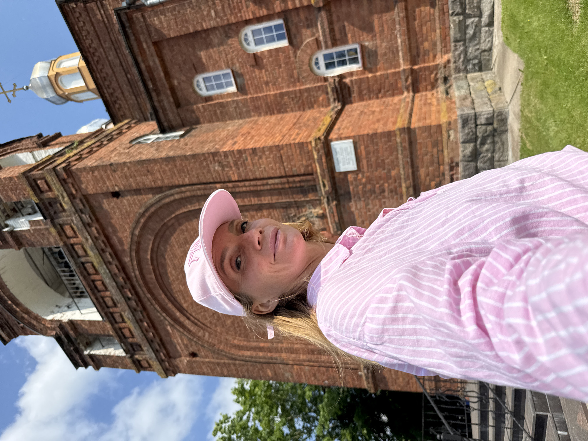 In an outdoor, low-angle, medium shot, a woman wearing a pink baseball cap and a pink and white striped button-down shirt is positioned in the foreground, facing right and slightly towards the camera. She has blonde hair and is looking at the camera with a slight smile. Behind her, a large, red brick building, identified as the Church of the Nativity of the Theotokos in Chudovo, Russia, dominates the background. The church features an arched entranceway, arched windows with white trim, and a silver dome topped with a cross. The sky is blue with scattered white clouds. To the left of the church, green trees are visible, and a set of stone steps with a black metal railing leads up to the building. The ground in front of the church is covered in green grass and stone.