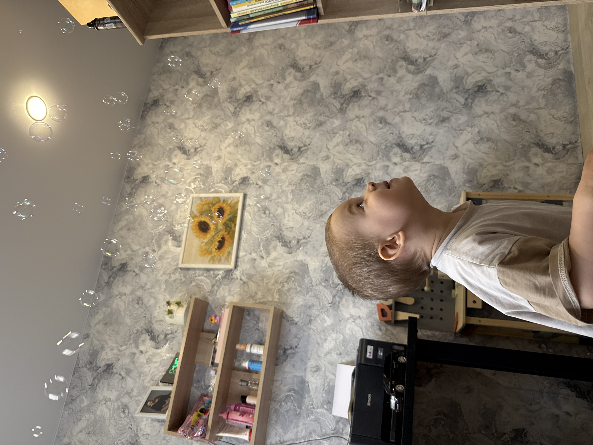 A child looks up at soap bubbles in a room. The room has a textured gray and white wall, a framed painting of sunflowers, a shelf with decorative items and storage, and a printer on a desk. The location is Rusanovo, Russia.