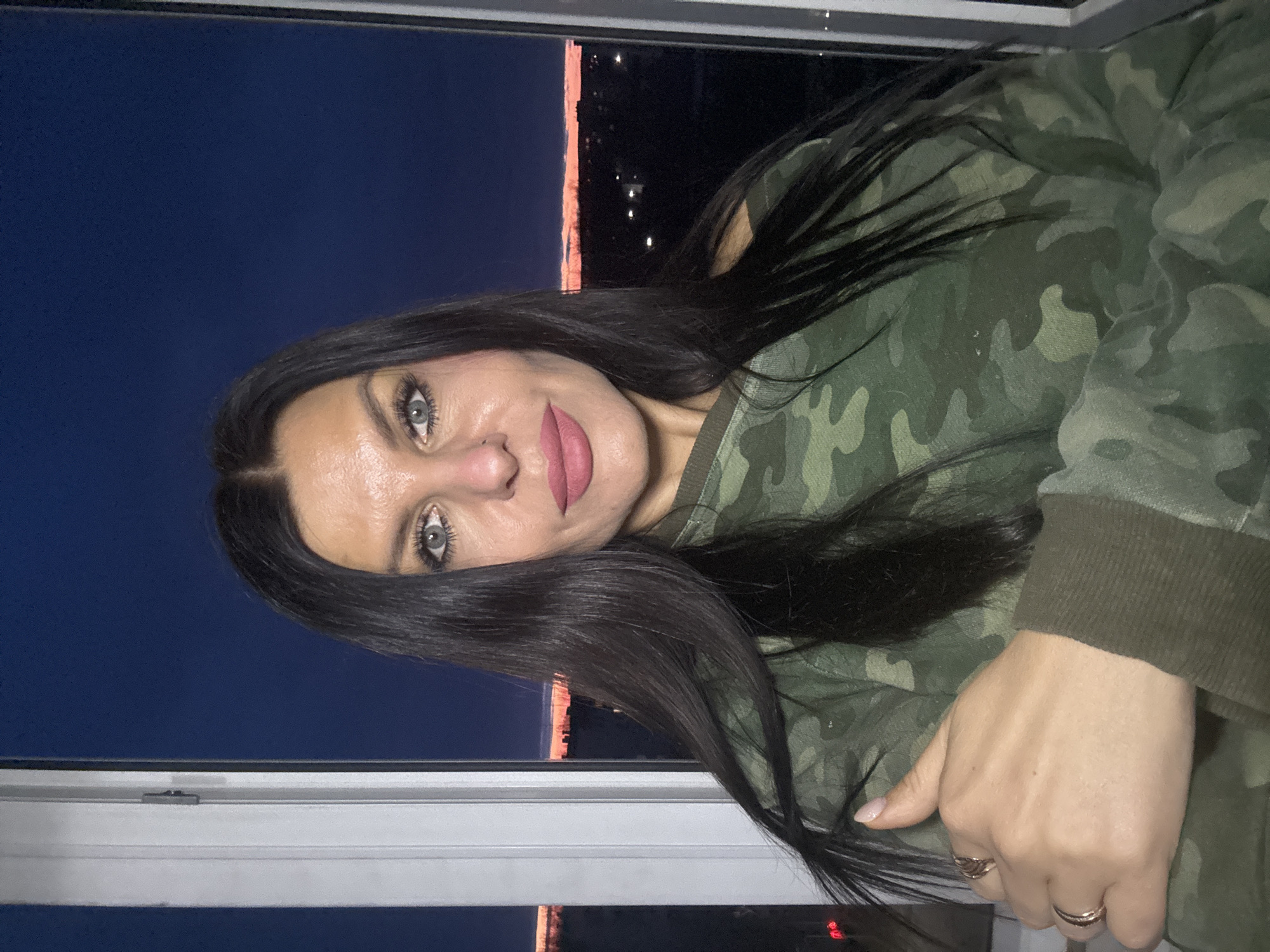 A medium close-up, horizontally oriented shot shows a woman posing for a photo indoors, looking directly at the viewer with a slight smile. She has long, dark hair, blue-gray eyes, and wears dark pink lipstick. Her skin appears smooth, and she has visible eyelashes.
She is wearing a green camouflage-patterned sweatshirt, which is visible on her upper body and arms. Her right hand is visible, resting near her side, adorned with two thin, possibly gold-toned, rings on two fingers.
The woman is positioned near a window, with a dark night sky visible outside. Through the window, faint, distant orange and white lights of a city or town illuminate the darkness, suggesting buildings or infrastructure in the distance. The window frame, predominantly white, is visible along the bottom and right side of the image, separating the indoor and outdoor scenes. The overall setting implies a nighttime view from an elevated position in Chaikovsky, Russia. There is no visible text in the image.