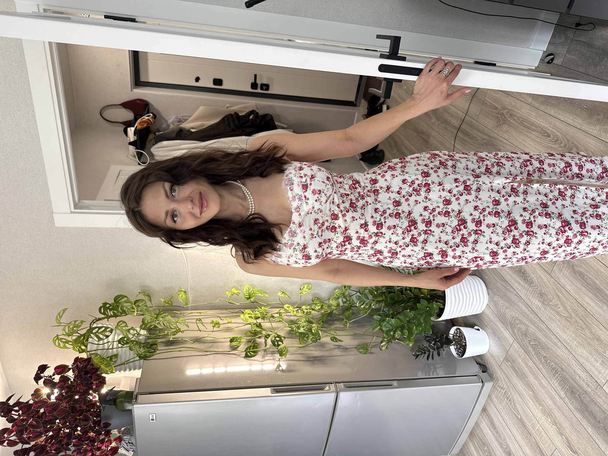 A woman in a floral dress stands in an apartment in Murino, Russia. She holds open a white door. The room beyond the door has clothing items scattered on the floor. To the woman's left is a silver refrigerator with various plants on top. The floor is light-colored wood.