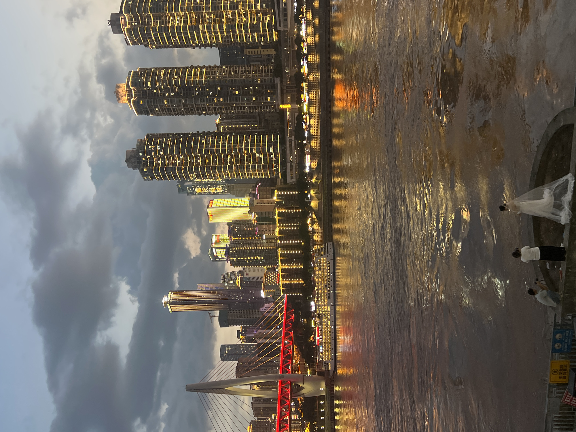 In Nan'an District, China, a river flows with reflections of city lights at dusk. A bridge with red accents spans the water, illuminated by the city's glow. Several high-rise buildings, adorned with golden lights, dominate the skyline. A large passenger boat is docked on the river, its windows brightly lit. On the riverbank in the foreground, a photographer kneels, taking a picture of a woman in a white wedding dress. The sky above is filled with dramatic, dark clouds.