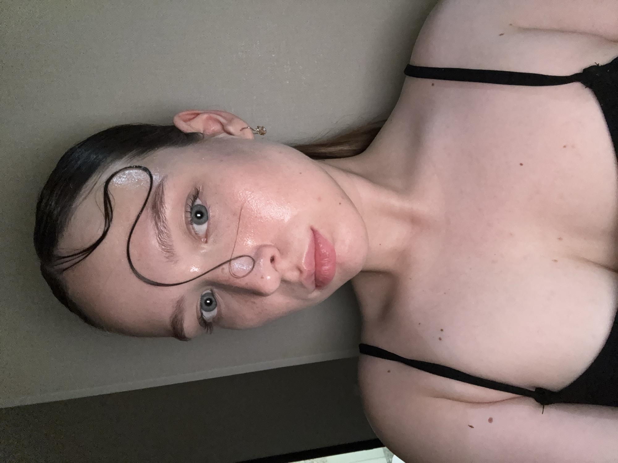 A young adult female is depicted in a close-up, frontal portrait, likely a selfie. She has light skin with visible freckles across her forehead and nose, and her face appears moist or glistening. Her eyes are blue-grey and directed towards the viewer, with a neutral expression. Her lips are naturally pigmented pink and slightly parted. Her dark hair is pulled back, but a single, wet, dark strand is styled in an elaborate, wavy pattern across her forehead, right temple, and down the bridge of her nose. She wears a black garment with thin spaghetti straps, revealing her shoulders and upper chest, where several small moles are visible. A small, simple metallic hoop earring is visible in her right ear. The background consists of a plain, light-colored wall, with a darker, shadowed vertical element visible on the left side of the frame.