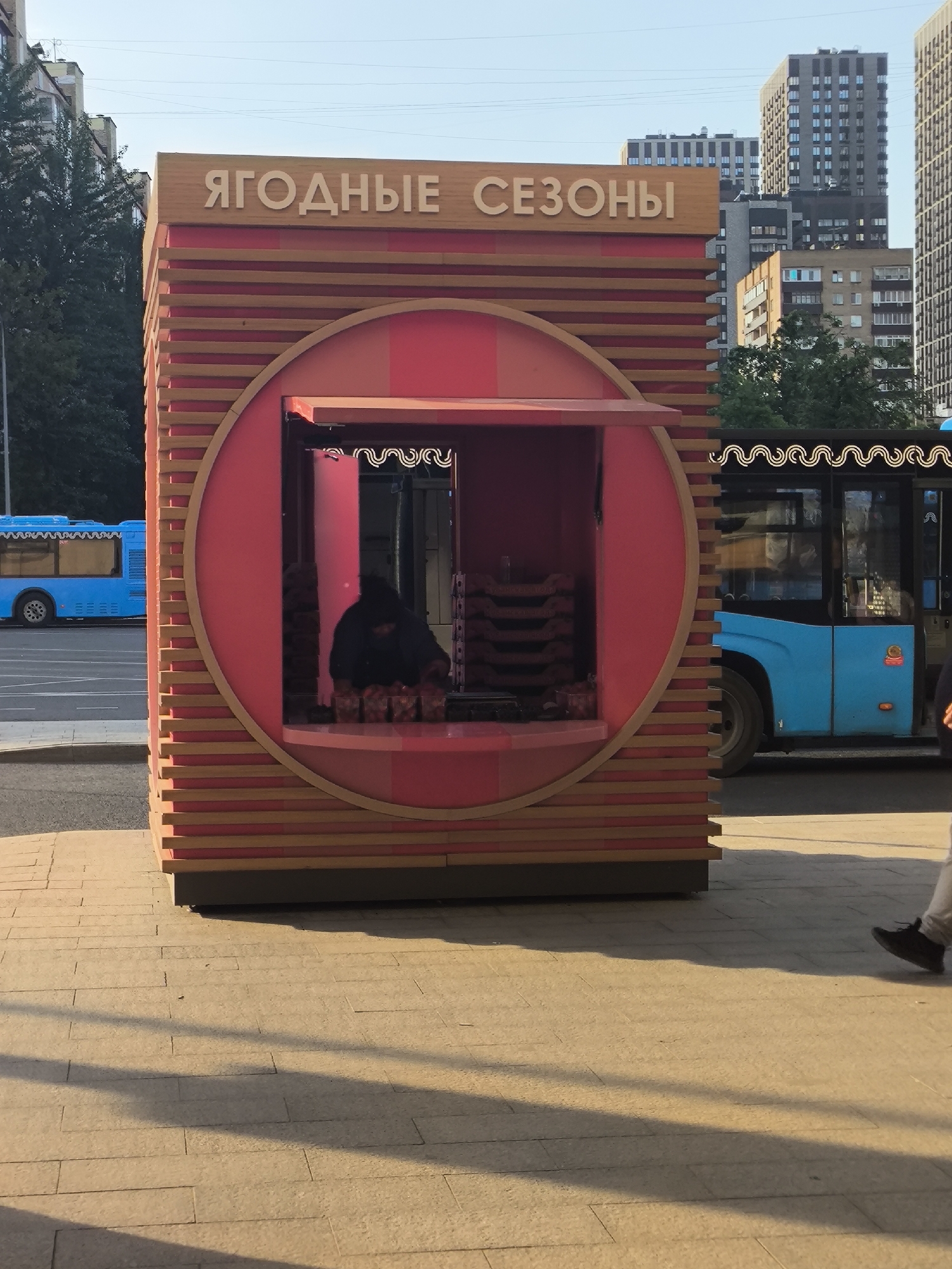 The photo captures a street scene in what appears to be an urban setting. In the foreground is a red and wooden kiosk or booth with a circular window-like opening, labeled "ЯГОДНЫЕ СЕЗОНЫ" (Yagodnye Sezony), which translates to "Berry Seasons". Inside the kiosk, a figure dressed in dark clothing is seen tending to what appears to be goods, possibly food items. A stack of containers with merchandise is also visible inside.
Behind the kiosk, partially visible on the left, is a blue bus. Another blue bus is partially visible on the right, with passengers inside and decorative wavy lines above the windows.
In the background, various buildings can be seen, including high-rise apartments, indicating that the location is likely a city or large town. The sidewalk in the foreground, made of rectangular paving stones, shows shadows cast by the setting sun. To the right, a person's legs and feet can be seen walking along the sidewalk.