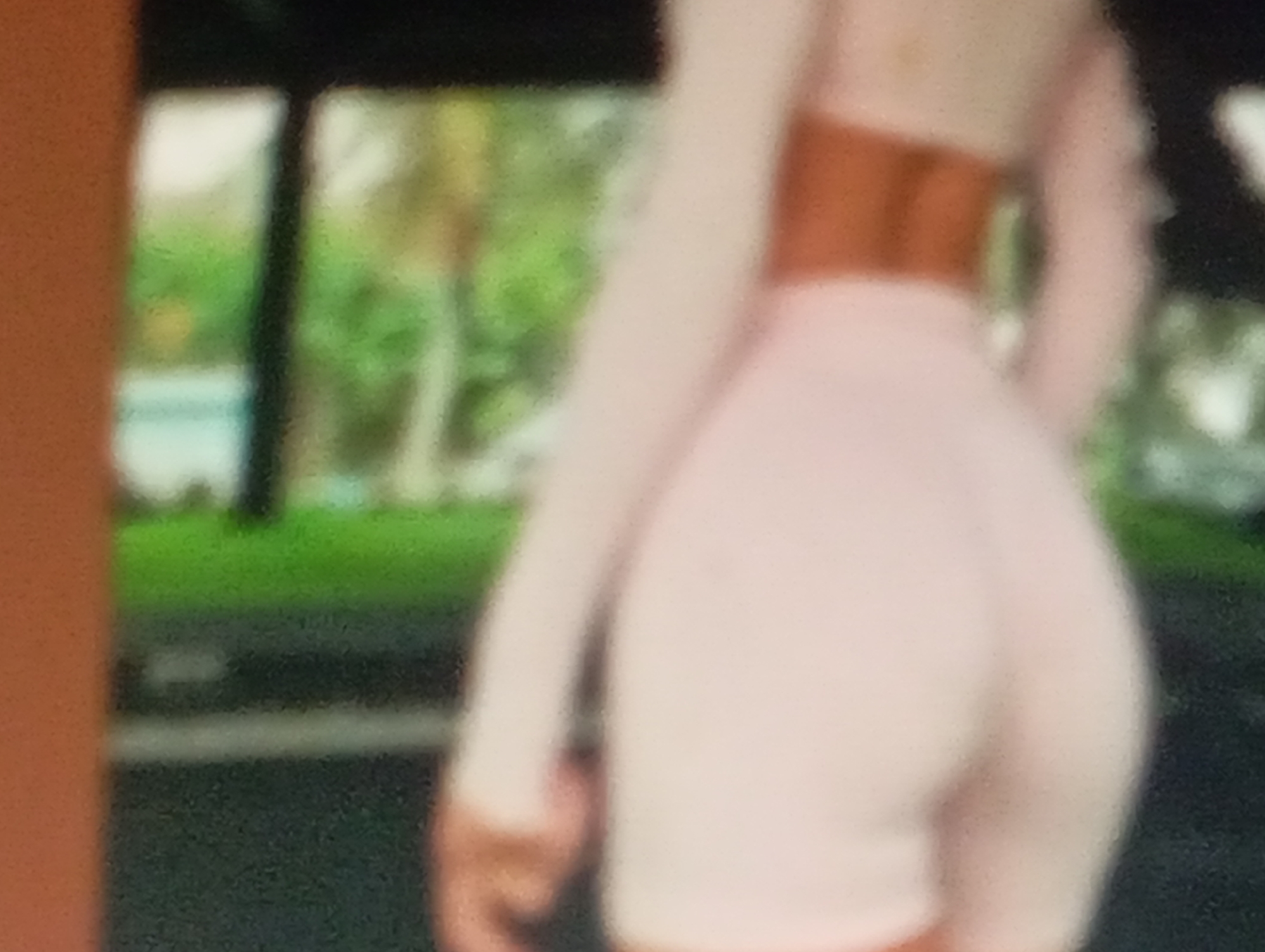 The image is a close-up, blurred photograph of a person wearing a light-colored long-sleeved crop top and matching shorts. The background suggests an outdoor setting with greenery and possibly a building structure with windows, hinting at a location like a park or an outdoor gym. No specific landmarks or details that would pinpoint the location to Sofiivska Borshchahivka, Ukraine are visible. The focus is on the person's attire and physique.