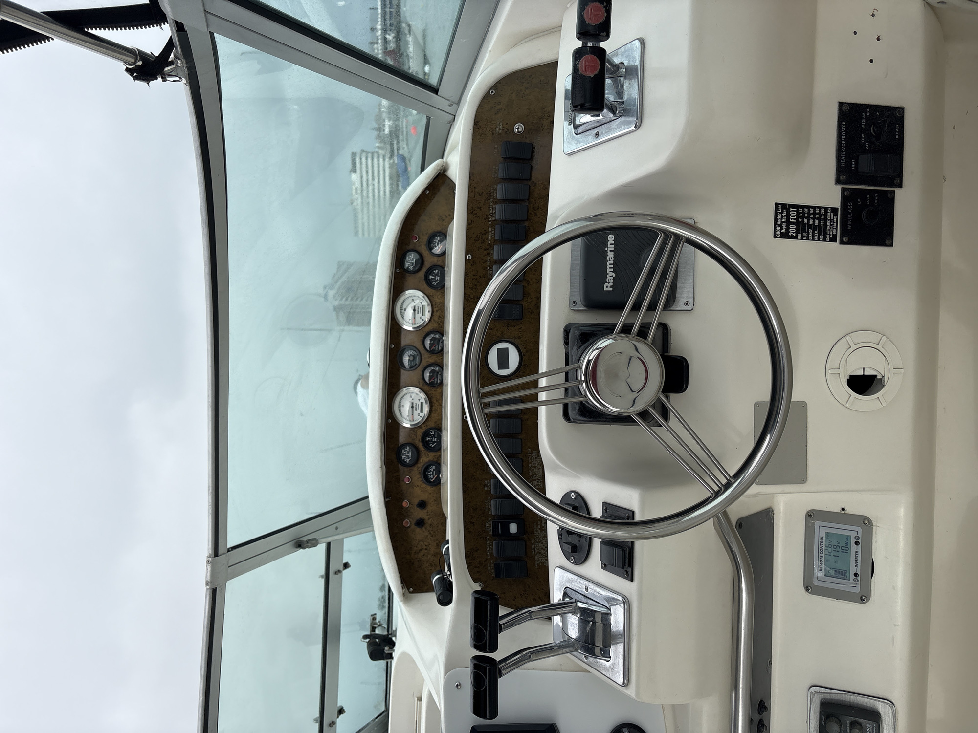 The image shows the interior helm of a boat, viewed from the perspective of the captain. The dashboard features a chrome steering wheel, multiple gauges, switches, levers, and a Raymarine device. The boat's windshield is visible, offering a view of a cloudy sky and a city skyline with buildings across the water. The scene is forensic and matter-of-fact, focusing on the details of the boat's interior and the external environment visible through the windshield. There are no people or interactions depicted in the image. The location is stated to be La Punta, Peru.