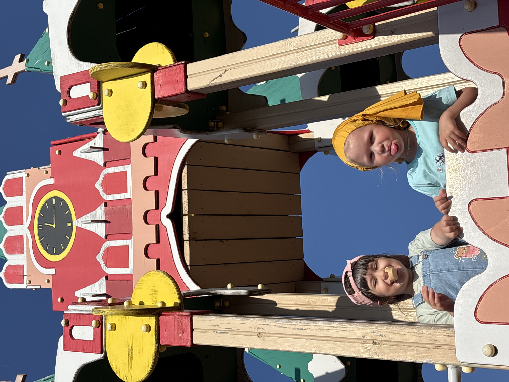 Two young girls are looking down from a colorful playground structure in what appears to be the city of Chaikovsky, Russia. The structure is made of painted wood and includes elements such as a clock face, stylized windows, and architectural details that give it a whimsical, storybook appearance. One girl is wearing a yellow headscarf and a light blue shirt, sticking her tongue out. The other girl is wearing a pink headband and denim overalls. She is holding a small, light-colored object to her mouth. The sky is blue and visible in the background. The location is determined based on the playground style and architecture, consistent with what is commonly seen in Chaikovsky.
