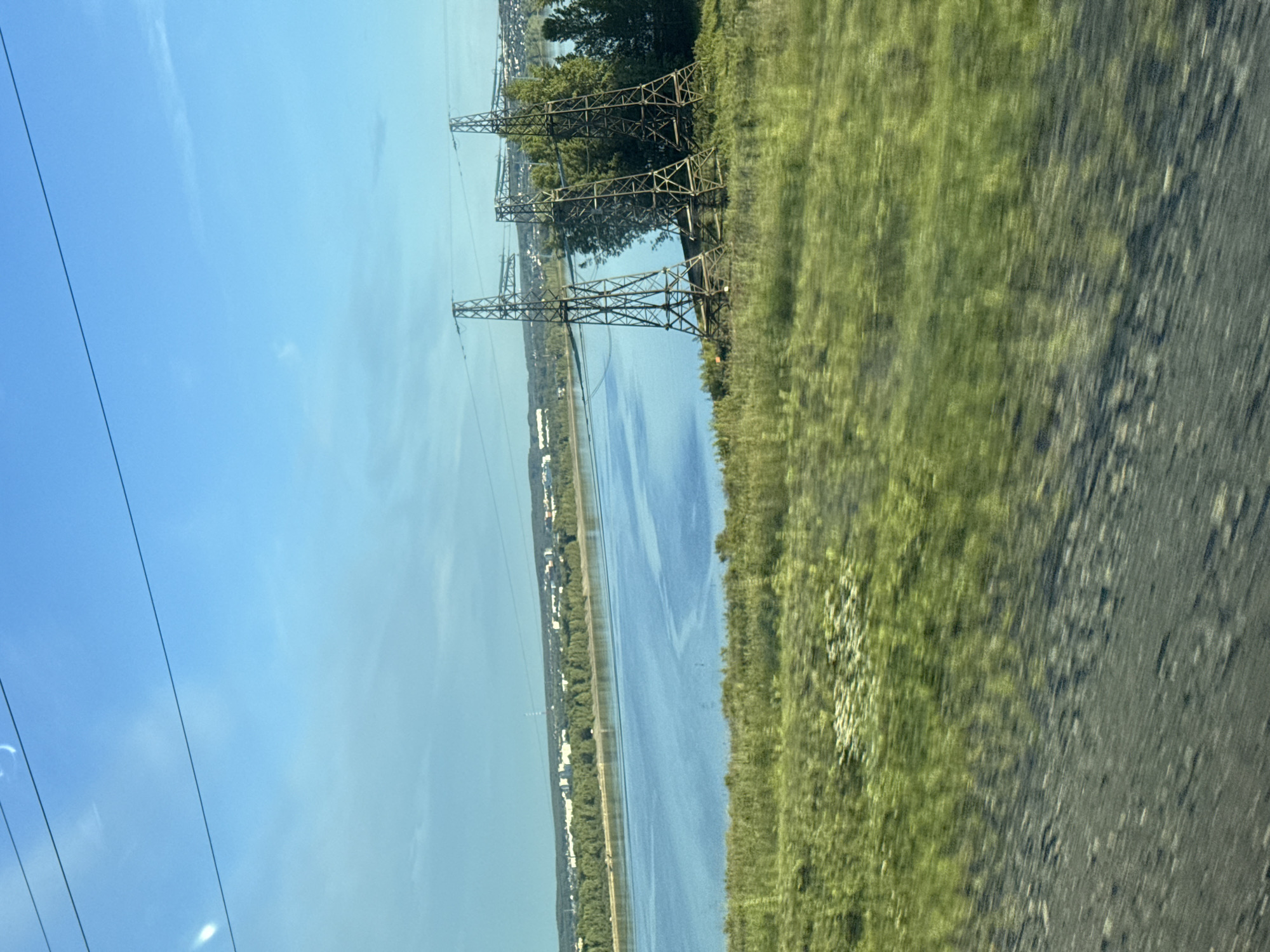 The image, taken from a moving vehicle in Chaikovsky, Russia, captures a vast outdoor scene under a clear blue sky. The scene depicts a wide body of water, likely a river or large lake, flanked by green landscapes and industrial infrastructure.
In the upper part of the frame, the sky is a bright, clear blue with only a few faint, wispy clouds visible. Several thin, dark power lines stretch across the sky, originating from large electricity pylons.
Dominating the midground are several towering metal electricity pylons, featuring intricate lattice structures. Two of these pylons are particularly prominent on the near side of the water, appearing to carry power lines across the expansive blue body. More pylons can be seen extending into the distance along the far bank.
The wide expanse of water is calm and reflects the clear sky, creating a serene blue surface. The near bank of the water is densely covered with lush green trees and foliage. On the distant shore, a continuous line of green trees is visible, behind which a scattering of white and light-colored buildings suggests a small town or settlement.
The immediate foreground on the right side of the image shows a patch of vibrant green grass and what appears to be a blurry road shoulder, with significant motion blur, indicating the photograph was captured from a fast-moving vehicle. The time of day appears to be bright daytime, likely late morning or afternoon, given the clear conditions and strong sunlight. No discernible text is visible in the image.