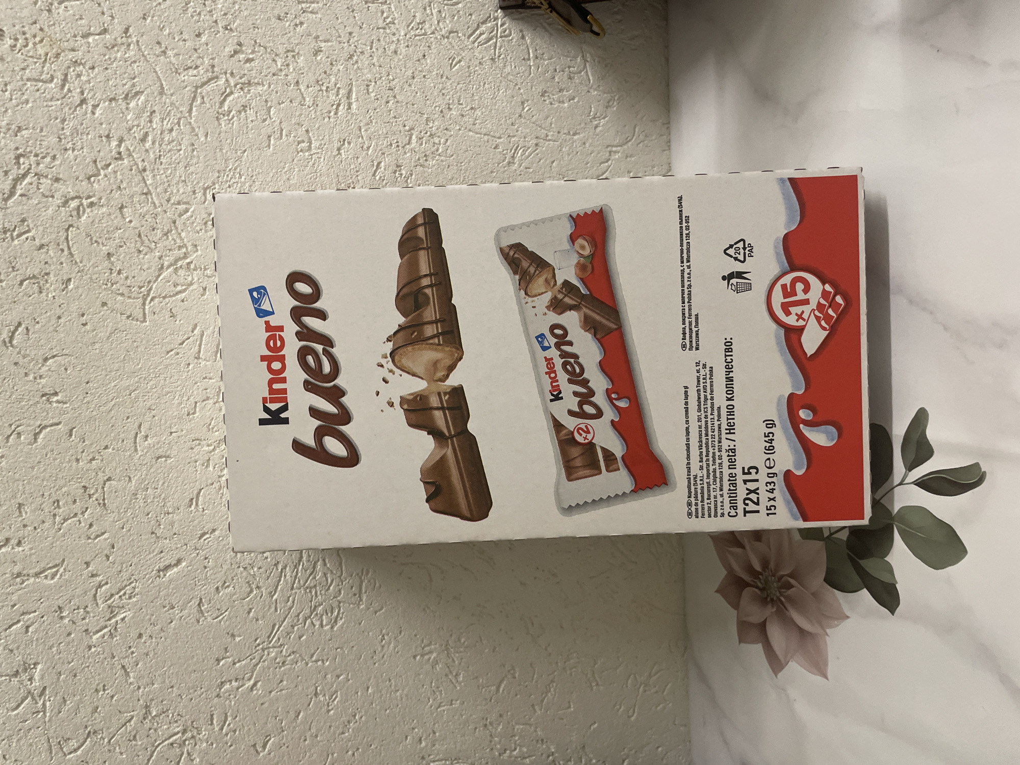 A slightly rotated eye-level close-up shot displays a box of Kinder Bueno chocolates on a white surface, likely a marble countertop. The box is white with the brand name "Kinder Bueno" in brown lettering on the left side. An image of a partially unwrapped Kinder Bueno bar is prominently displayed on the box, alongside the phrase "T2x15" indicating the quantity and weight (15 x 43 g = 645 g). To the bottom right of the box, a pale pink flower and some greenery are partially visible. The backdrop consists of a textured off-white wall. The location is not Sofia, Bulgaria based on the text on the box. The text indicates the product is manufactured in Poland and imported to Romania and Moldova.