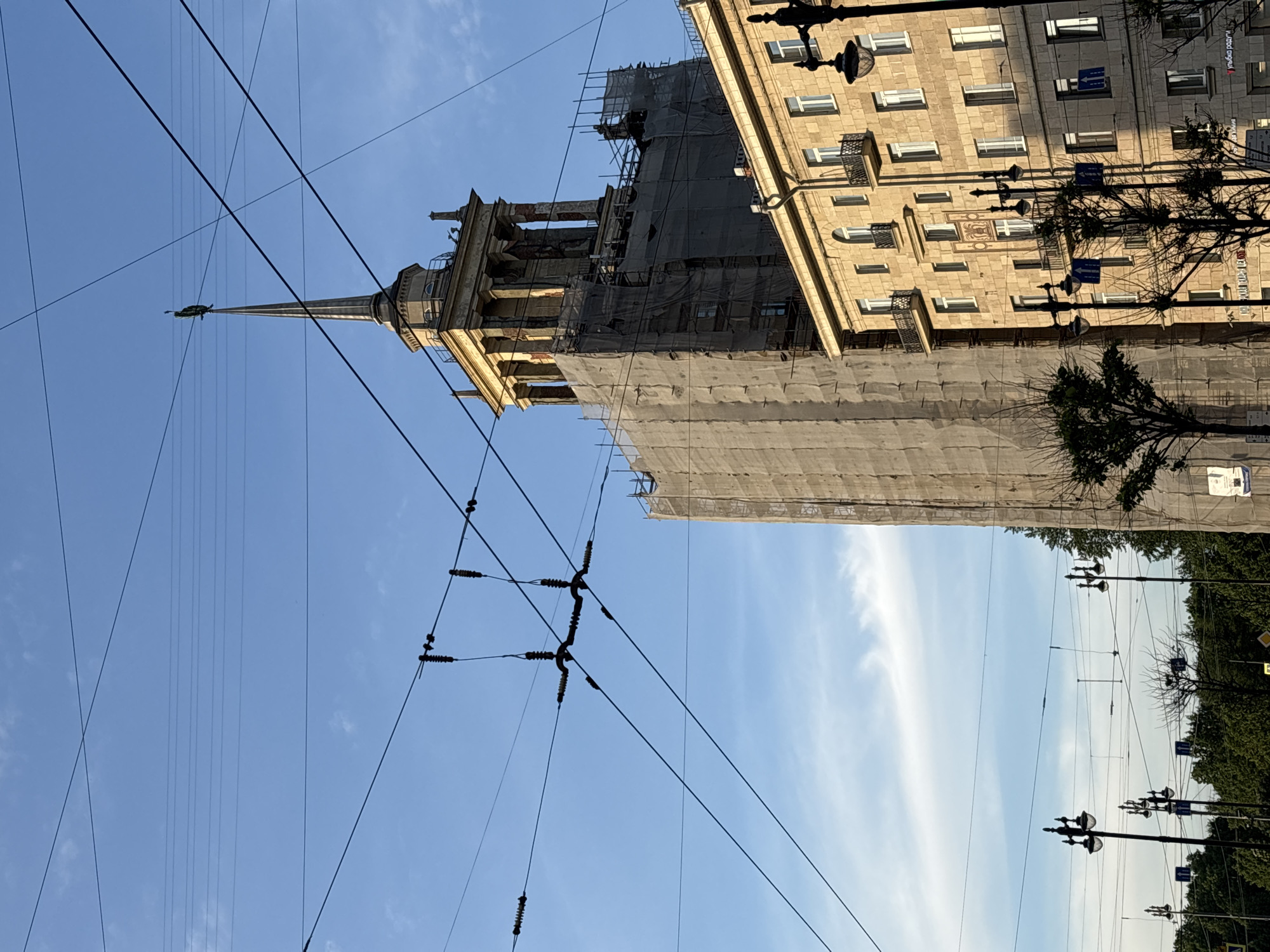 The image depicts a tall, imposing building undergoing significant construction or renovation, set against a clear blue sky with sparse white clouds. The perspective is looking upwards, showcasing the building's height and the crisscrossing network of overhead power lines.
The central subject is a multi-story building, likely an example of Stalinist Neoclassical architecture given the context of Saint Petersburg. Its upper sections feature ornate classical elements, including columns and decorative cornices, culminating in a slender spire. At the very peak of the spire, a small, dark, bird-like ornament or weather vane is visible. A substantial portion of this building's facade is covered in scaffolding and light-colored netting or tarps, indicating active renovation work. Below the ornate top, the main body of the building is largely obscured by these coverings.
To the right of the central building, an adjacent, lighter-colored stone or stucco building is visible. This building has multiple floors with numerous rectangular windows, some featuring small, dark, wrought-iron balconies. Several black street lamps with multiple heads are positioned in front of this building. Shadows cast on the building suggest bright daylight conditions.
A dense array of dark electrical wires and power lines, some with insulators, dominate the foreground and crisscross the sky, indicative of urban infrastructure, possibly for public transport like trams or trolleybuses. These lines create a geometric pattern against the clear sky.
Visible text includes two blue directional signs with white arrows on the street lamp poles in front of the adjacent building, one pointing left, the other right. Cyrillic text is discernable on at least one of these signs, though too small to fully decipher. Another faint Cyrillic text, possibly "УЛ. ЛЕНИНА" (Lenin Street), can be vaguely seen on a banner or sign on the adjacent building. The presence of these architectural styles and Cyrillic text aligns with the given location context of Saint Petersburg, Russia. The overall mood is bright and active, reflecting ongoing urban development or maintenance.