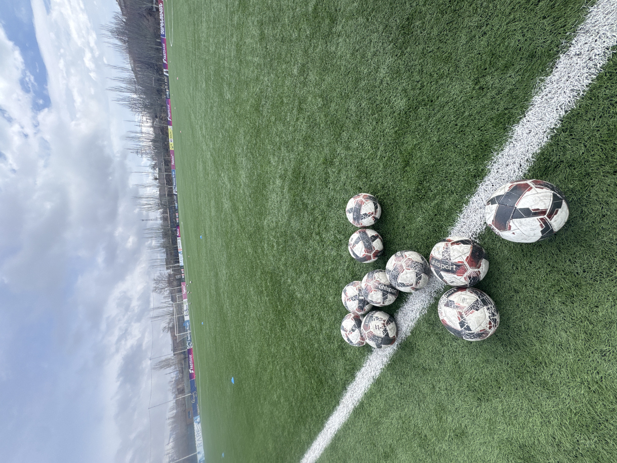 The image shows a corner of a green artificial turf soccer field in Charentsavan, Armenia. A white painted line marks the field boundary. There are ten soccer balls clustered near the corner. One ball has the word "uhlsport" printed on it. Behind the field, a barrier displays advertisements, including the words "FASTBANK" and "fanshop.fe.am". Bare trees stand further behind the barrier. A cloudy sky is visible.