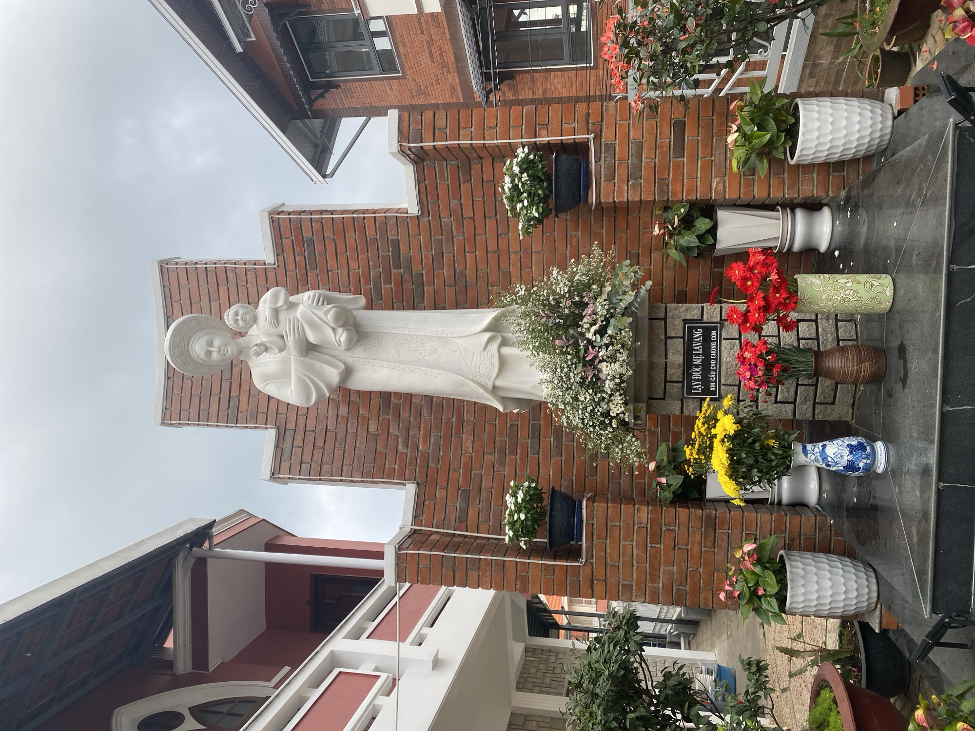 The image displays an outdoor devotional scene in Lâm Viên - Đà Lạt, Vietnam, featuring a prominent white marble statue of the Virgin Mary, identified as "Our Lady of La Vang," holding the Child Jesus. The stylized statue, depicting the figures in traditional Vietnamese-inspired garments and headwear, stands on a dark base against a reddish-brown brick wall.
Numerous potted plants and fresh floral arrangements surround the statue. A large cluster of white and light pink flowers adorns the base, while smaller arrangements of red, yellow, and white flowers, along with various green plants, are artfully placed in different-shaped pots (white textured, plain white, green patterned, and brown) on the dark grey tiled steps in front of the brick wall. Hanging baskets with white flowers are also mounted on the brickwork.
In the background, parts of two residential buildings are visible under an overcast sky. One building behind the statue features red bricks, dark-framed windows, and a dark tiled roof. To the left, a red and white building with a tiled roof and white architectural elements is partially seen.
A black sign with white Vietnamese text is visible next to the statue. The text reads: "LẠY ĐỨC MẸ LAVANG" which translates to "Oh Our Lady of La Vang," and "XIN CẦU CHO CHÚNG CON," meaning "Please pray for us." The cloudy weather provides soft, diffused lighting, contributing to the tranquil and reverent atmosphere of this beautifully maintained religious display.