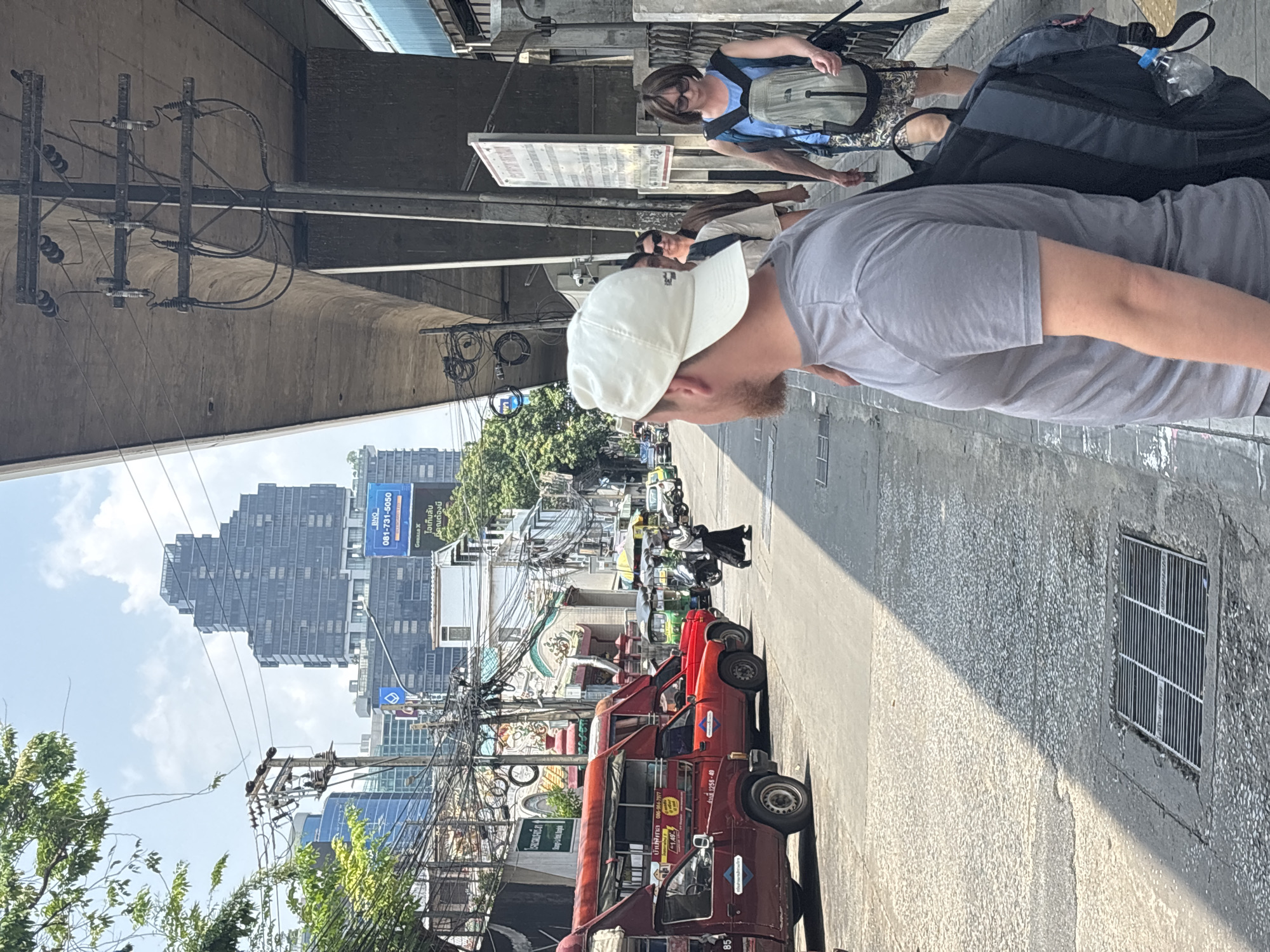 The image shows a street scene in Bangkok, Thailand. A man with a white baseball cap and gray shirt is in the foreground. Behind him are other people, including a woman wearing a blue shirt and backpack. The street is lined with red tuk-tuks. Power lines crisscross overhead, and there is a large building in the distance. A sign displays phone numbers.