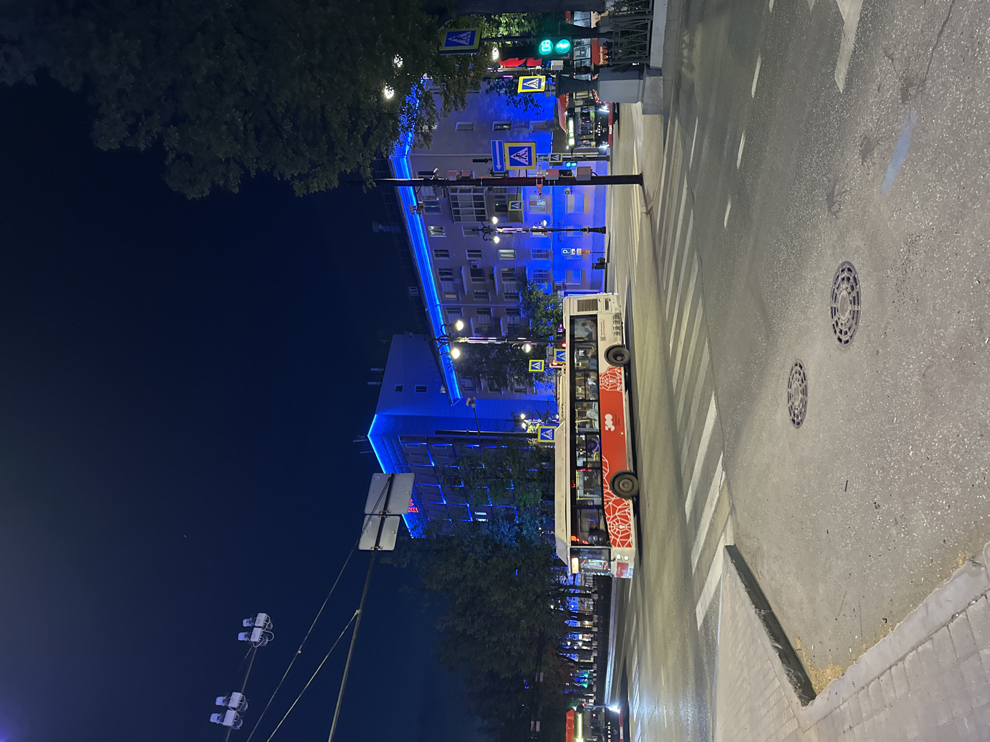 The image is taken at night in Perm, Russia, showcasing a street scene. The street is wet, reflecting the city lights. A bus with a red and white design is stopped at a zebra crossing. Pedestrian crossing signs are visible on the street. The building across the street is lit with blue lights, emphasizing its architecture. Street lamps illuminate the scene. The sky is dark.