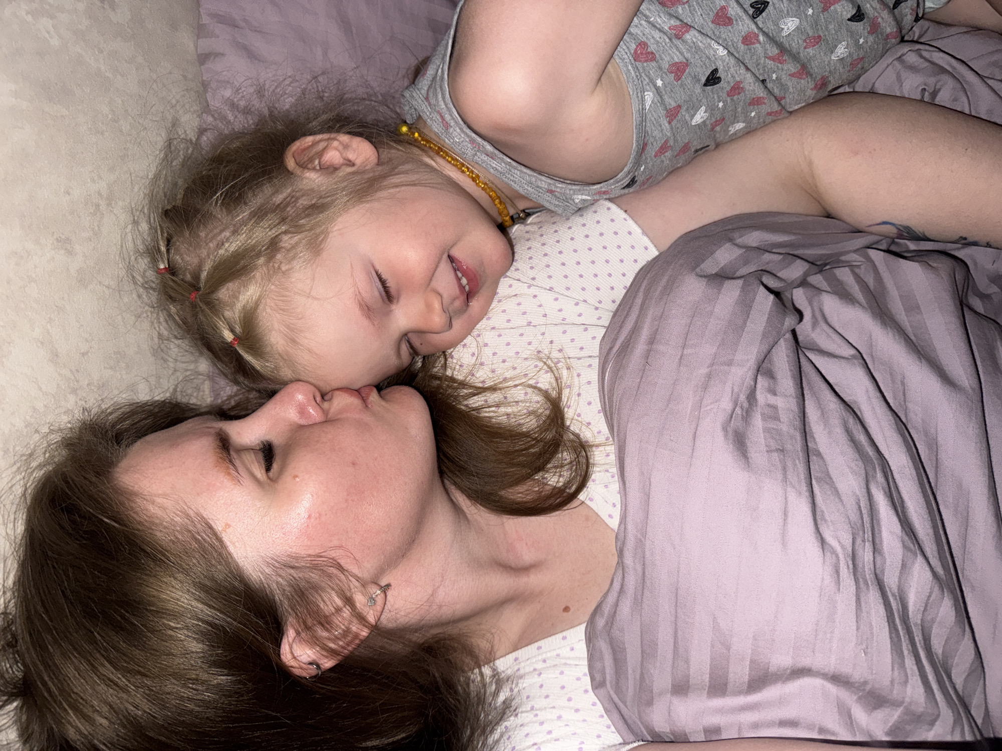 An intimate portrait captures an adult female and a young child, likely a mother and daughter, lying side-by-side on a bed in Gornyi (Dvurechenskoe S/P), Russia. The adult, with shoulder-length brown hair, is positioned on the left, gently kissing the child's forehead or temple. She wears a light-colored top with subtle purple dots and small earrings. The child, with light blonde hair styled in multiple small pigtails secured with red elastics, lies on the right, smiling serenely with closed eyes. She is wearing a grey t-shirt adorned with pink and white heart patterns and a yellow beaded necklace.
Both subjects are partially covered by bedding; a white sheet with light purple dots beneath them, and a striped purple comforter over parts of their bodies. The soft, dim lighting suggests either evening, early morning, or a low-light indoor setting. The scene conveys a profound sense of tenderness, affection, and peaceful contentment between the two individuals. There is no visible text in the image.
