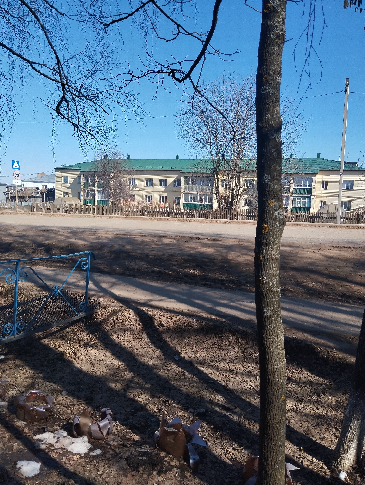 The image depicts a street scene in Krasnogorskoye, Russia. A two-story building with a green roof and yellow walls stands across the street, behind a wooden fence. Bare trees are present, with branches obscuring parts of the sky. A blue metal gate is visible on the left. The foreground shows patches of dirt, leaves, and remnants of snow. Some cylindrical metal objects, potentially discarded items or decorations, are also present near the gate. Shadows from the trees are cast across the ground. A speed limit sign is visible on the left.