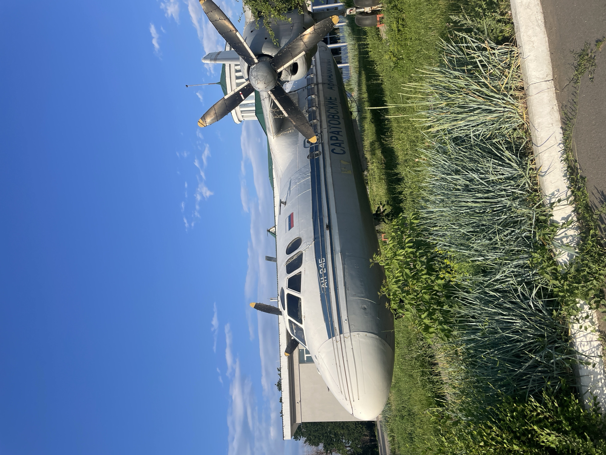 An Antonov An-24 turboprop aircraft, painted white with a blue stripe along its fuselage and "SARATOVSKIYE AVIA (" Saratov Airlines") written in Cyrillic, is parked on a grassy area in front of a low-rise building with a green roof. The aircraft has a prominent nose, a cockpit with multiple windows, and a four-bladed propeller with yellow tips. The sky is blue with scattered white clouds. The aircraft appears to be a static display, possibly at a museum or an airport in Saratov, Russia. The foreground is filled with overgrown grass and bushes.