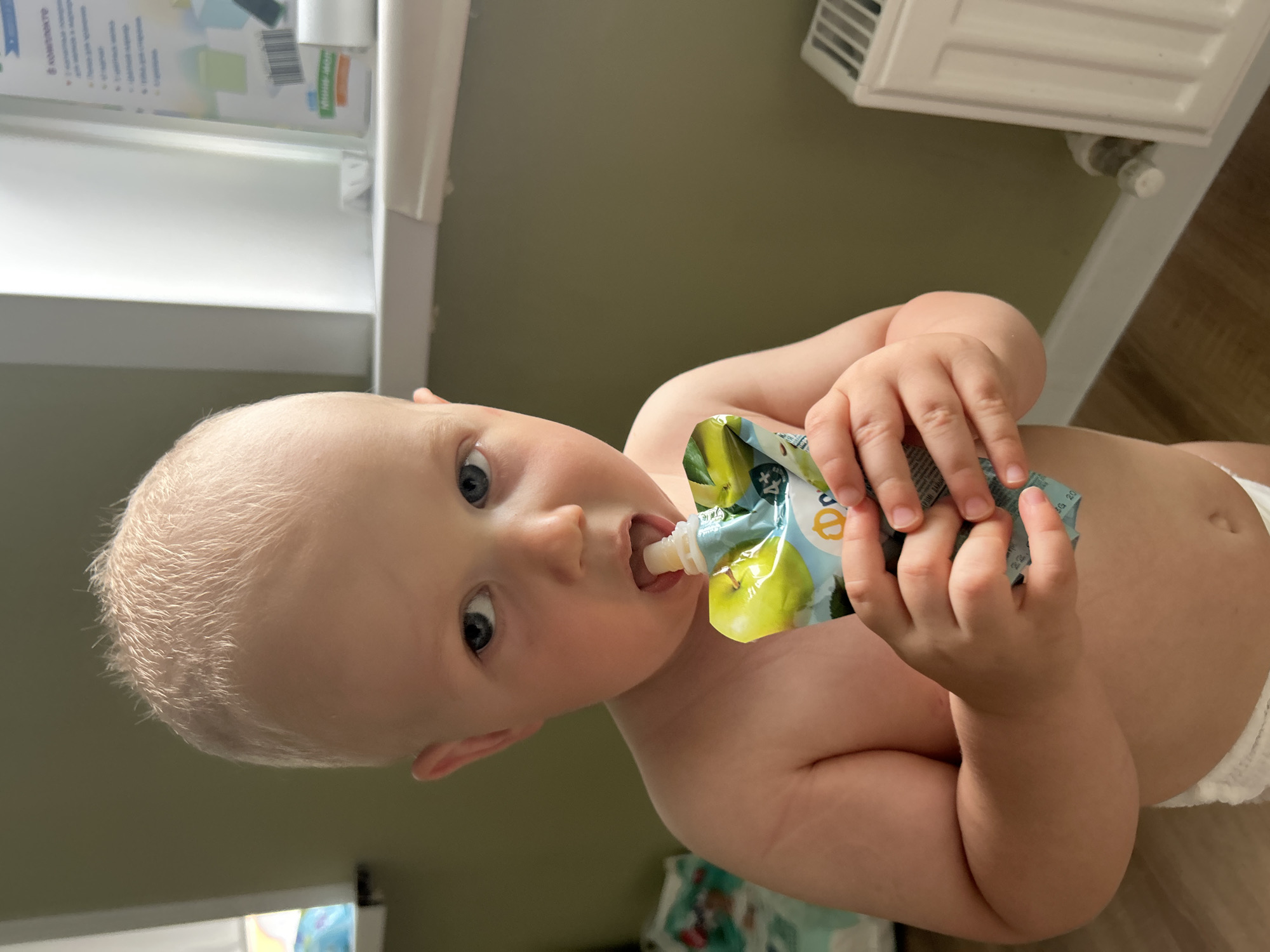 A toddler with blond hair and blue eyes is holding a pouch of baby food and putting the nozzle into their mouth. The child is wearing a diaper. The background is a wall of an olive green color, with a white windowsill on the right side of the image. There is a white radiator visible on the right side of the image, and a portion of a baby care product box is visible on the windowsill. The floor is made of wood. The image appears to have been taken indoors, in a residential setting. There is no information to suggest the location is in Kaluga, Russia.