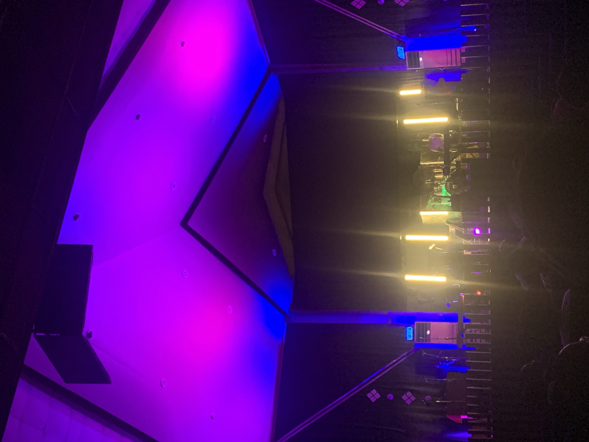 The image shows an indoor scene at a concert or event, likely a music performance. The left side of the image features a large, angular structure illuminated with purple and blue light, possibly part of the stage design. The right side displays a series of scaffolding or platforms with yellow and blue lights, possibly supporting equipment or serving as part of the stage. There are silhouettes of people in the lower right corner, indicating an audience. The overall lighting is dark with vibrant colors. There is no information available that indicates the location is in the city of Dar es Salaam, Tanzania.