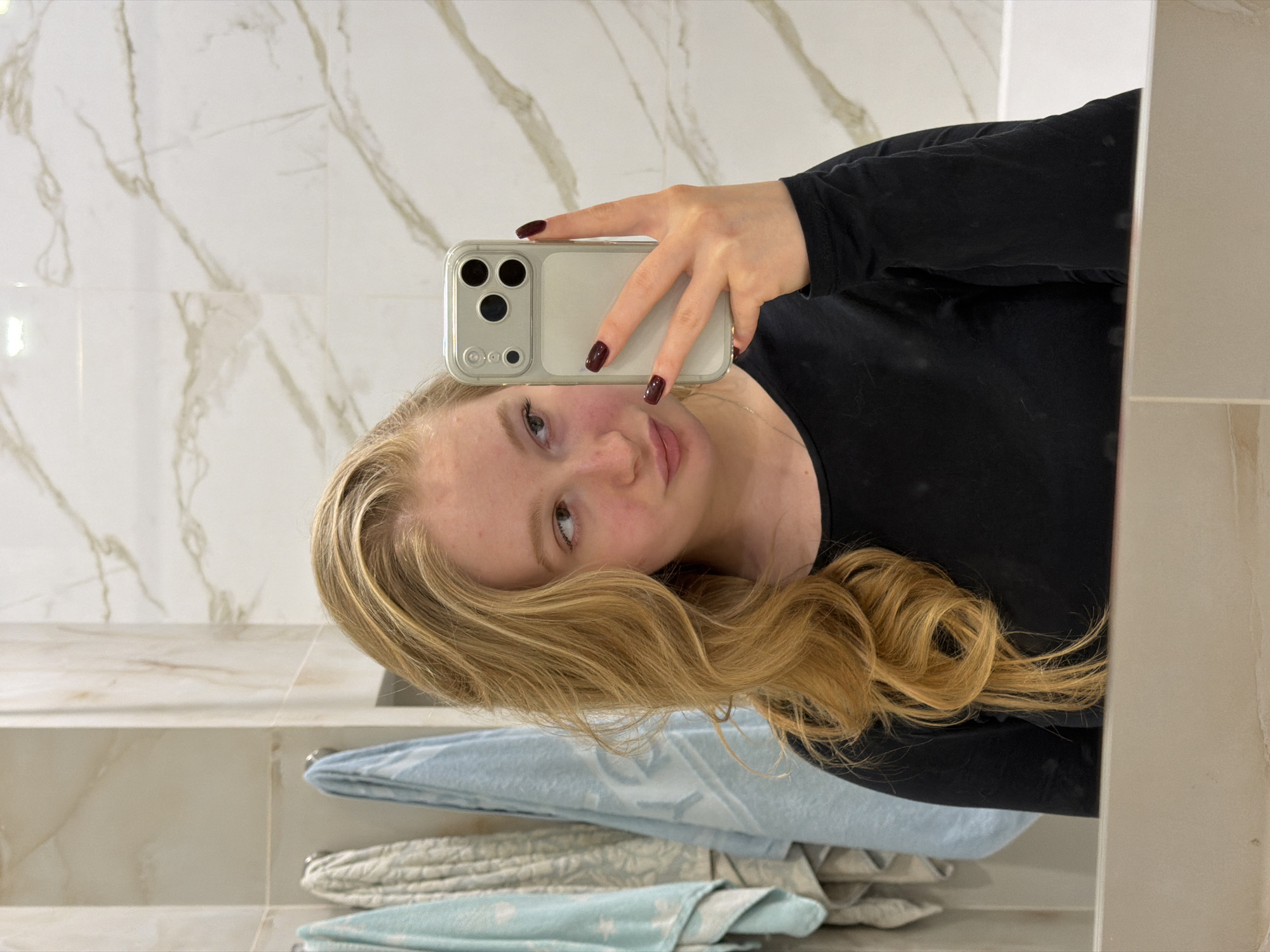 A fair-skinned, blonde woman is taking a selfie in a bathroom. She is wearing a black, long-sleeved shirt and holding a silver smartphone. Her fingernails are painted dark red. The background consists of white marble-patterned tiles on the wall and a stack of folded towels on a shelf below the mirror. The scene appears to be indoors in a bathroom in an unknown location, but is specified as being in Rossiyskiy, Russia.