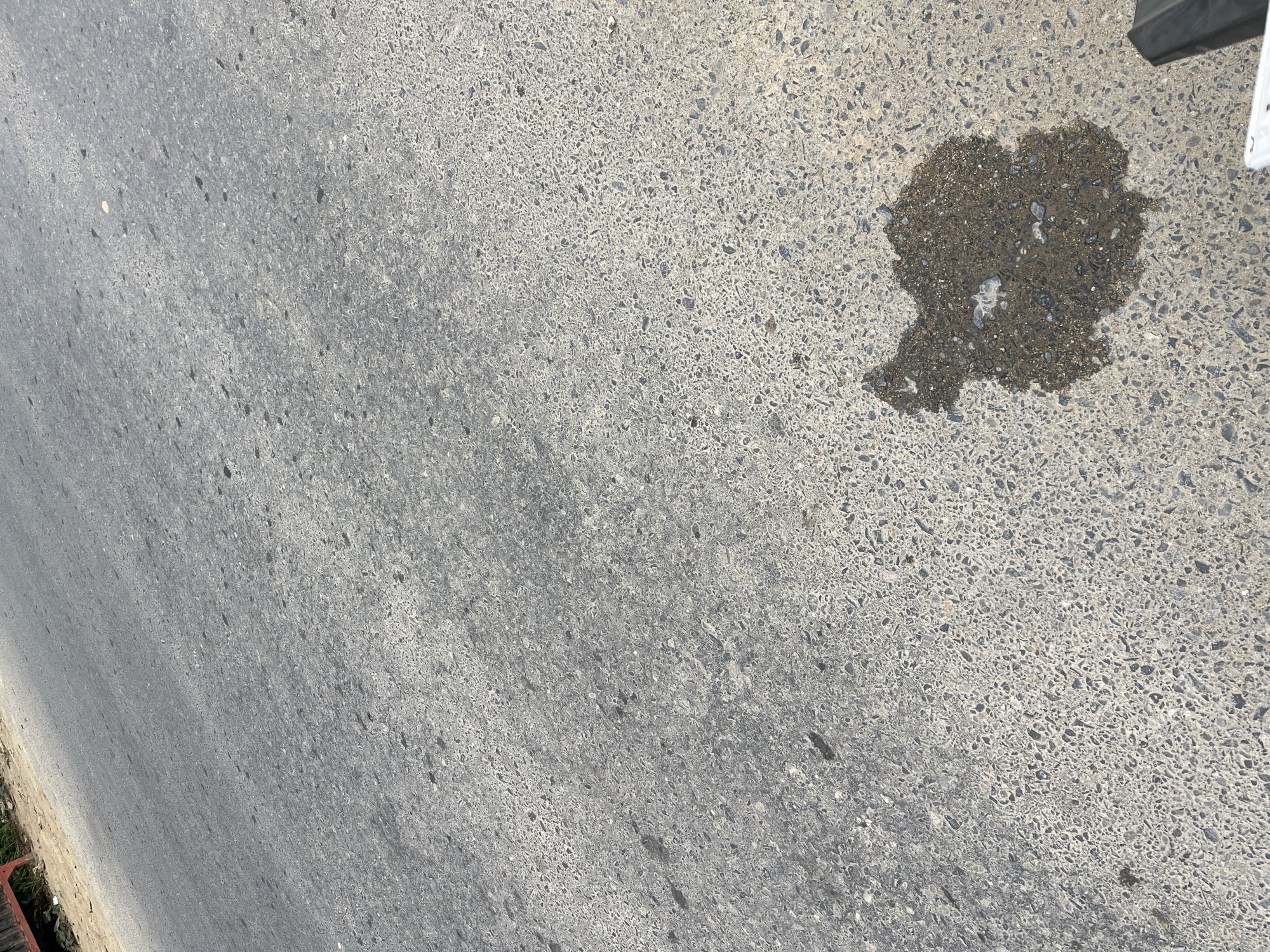 An overhead, slightly angled, medium shot captures a patch of asphalt road in Simpang Empat, Indonesia. The road surface is a coarse, grey aggregate with visible small stones and darker specks. In the lower right quadrant, a dark, irregular shaped puddle of water, appearing slightly murky with some debris, rests on the asphalt. The top left portion of the image shows the edge of the road where it meets a dirt embankment with some discarded materials. The lighting suggests it is daytime, casting subtle shadows on the road surface. There are no people or vehicles prominently featured in the scene, and the focus is on the road and the puddle.