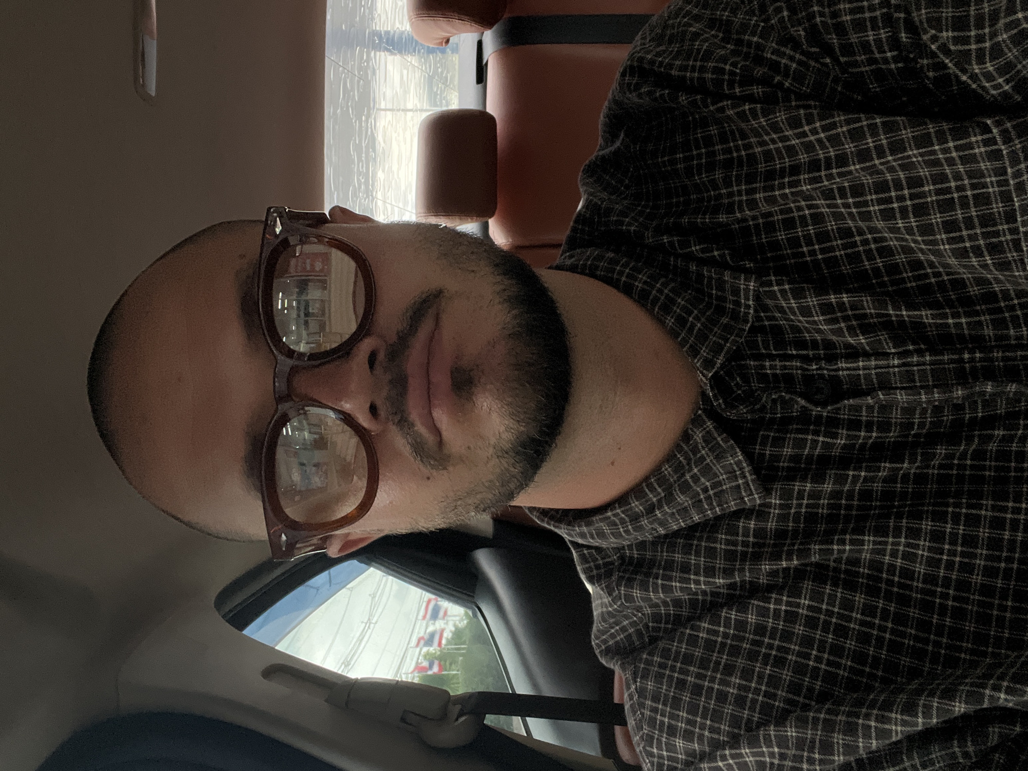 A medium shot of a man with a shaved head and beard wearing brown-rimmed glasses and a plaid shirt is taken inside a vehicle. The man is looking directly at the camera. The interior of the car, with its tan leather seats, is visible behind him. Outside the car window, several Thai flags are flying, and a building is reflected in the man's glasses. The background is blurred, suggesting the vehicle is in motion.