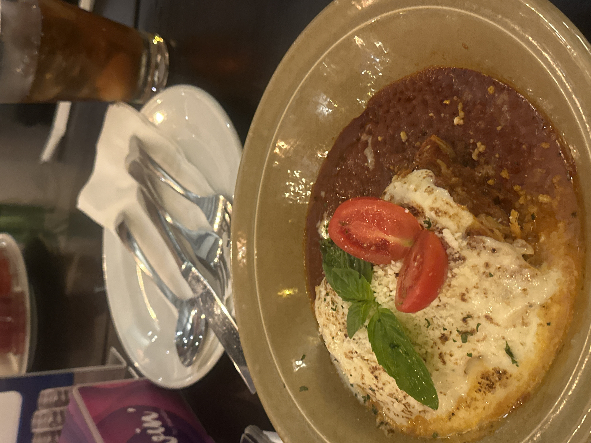 A close-up shot captures a dish of pasta with marinara sauce and melted cheese, garnished with fresh basil and sliced cherry tomatoes. The food is served in a shallow, light brown ceramic bowl. In the background, a white plate holds silverware, and a partially obscured beverage glass filled with iced tea is visible. A purple and pink napkin dispenser with text is present on the left. The setting appears to be a restaurant table in Denpasar, Indonesia, indicated by the ambient lighting and table setting typical of dining establishments in the area.