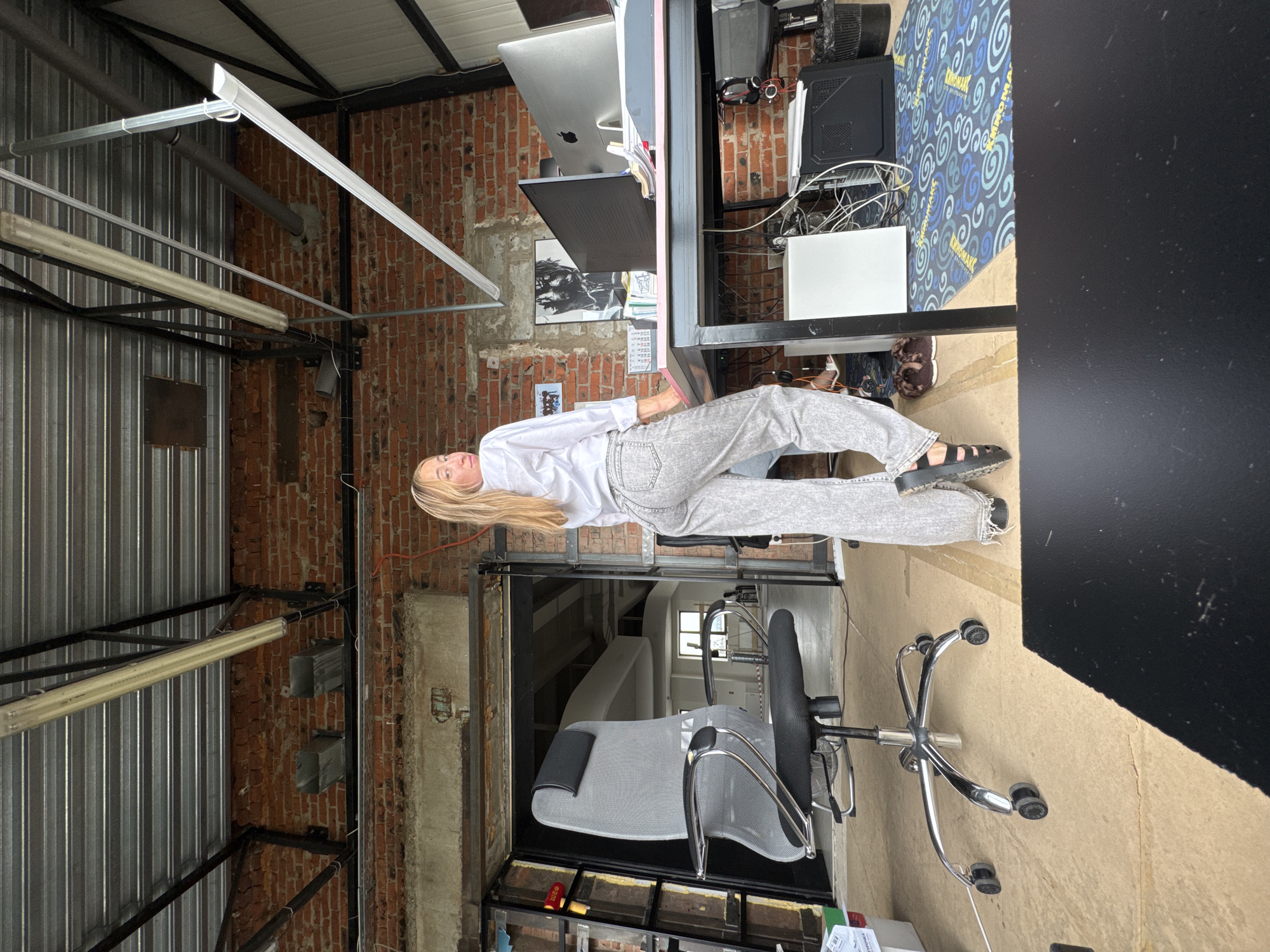 A young woman with blonde hair, wearing a white button-down shirt and light-wash denim pants, stands facing away from the camera and looking over her shoulder. She is positioned in an open-plan office space with exposed brick walls, a corrugated metal ceiling, and industrial-style lighting fixtures. To her left is a grey office chair with a mesh back. To her right, a desk is set up with multiple computer monitors, a computer tower, and various office supplies. The floor is covered with plywood and a patterned rug. The overall impression is a modern, somewhat unfinished office environment, possibly a workspace or a loft-style apartment. The image does not contain any specific details that would indicate its location within Chaikovsky, Russia.