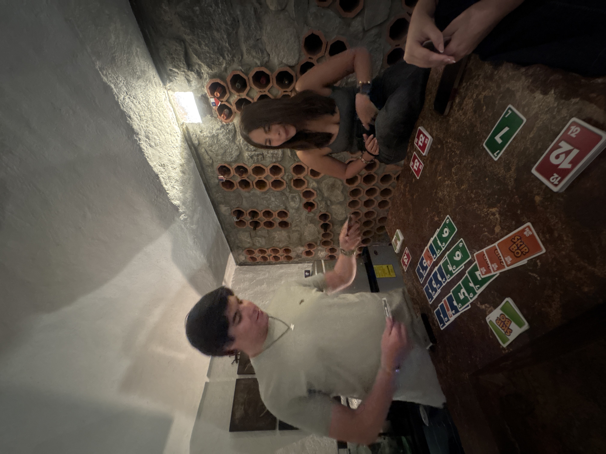 Two people are playing a card game at a table in a room with stone walls that have decorative clay pots embedded in them. One person is standing and appears to be in the process of playing a card, while the other is sitting at the table and watching. The cards are scattered across the table. The photo was taken in San Bernardino Tlaxcalancingo, Mexico.