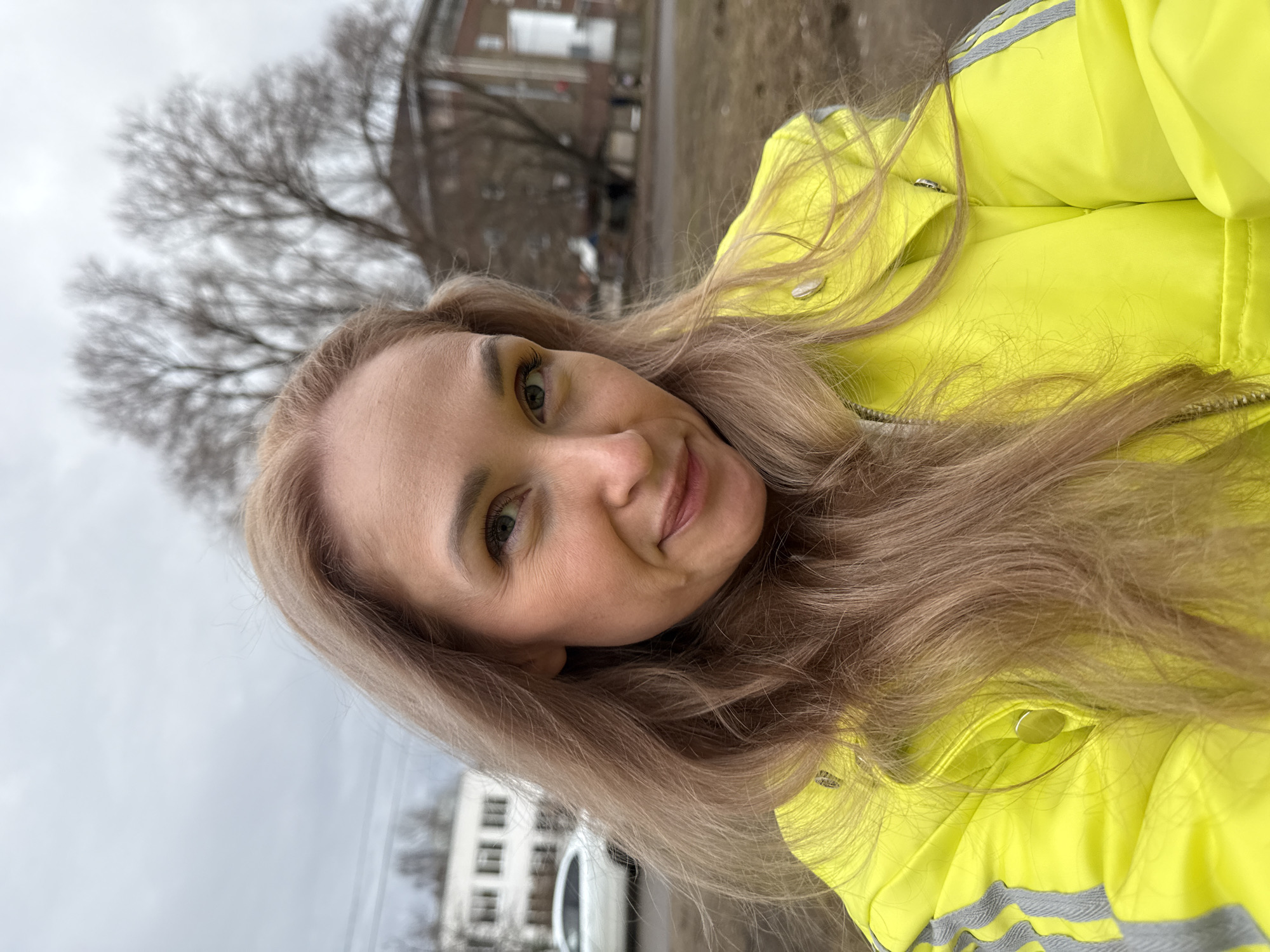 A woman with long blonde hair is posing outdoors, wearing a bright yellow jacket with silver reflective stripes on the shoulder area. She is smiling slightly, looking towards the camera. In the background, blurred, are buildings typical of an urban environment, with a gray, overcast sky and bare trees suggesting late autumn or early spring. A car is partially visible in the lower left corner. While specifics are lacking in the image to determine the location with certainty, the urban and architectural elements are consistent with the style found in Chaikovsky, Russia. The focus of the image is on the woman, and the background is secondary.