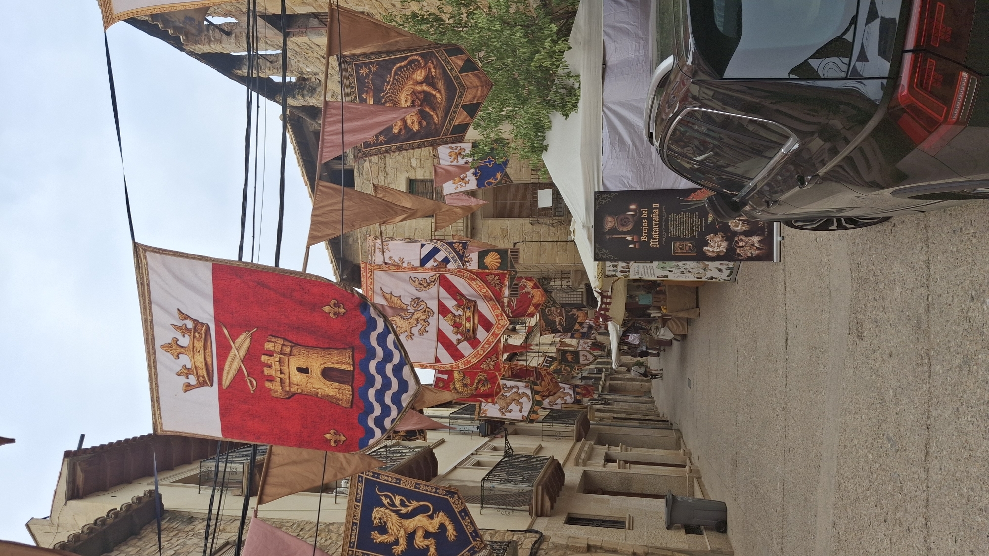 In the city of La Fresneda, Spain, a street is adorned with numerous medieval-style banners, hanging from wires stretched across the narrow, paved roadway. The banners feature heraldic symbols, including crowns, castles, lions, and mythical creatures, rendered in vibrant colors. The architecture of the surrounding buildings is predominantly stone, with balconies and arched windows, indicative of historical construction. On the right side of the street, white tents suggest a market or event is taking place, with a black SUV parked alongside. A banner in front of the tents advertises "Brujas del Matarraña II." A few people are visible in the distance, walking down the street, one accompanied by a dog. The sky is overcast, casting a soft light on the scene.